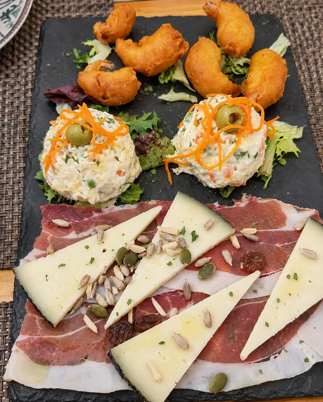 A slate board with fried fritters, two quenelles of potato salad topped with an olive and grated carrot, and triangles of cured cheese over slices of ham with seeds and raisins.