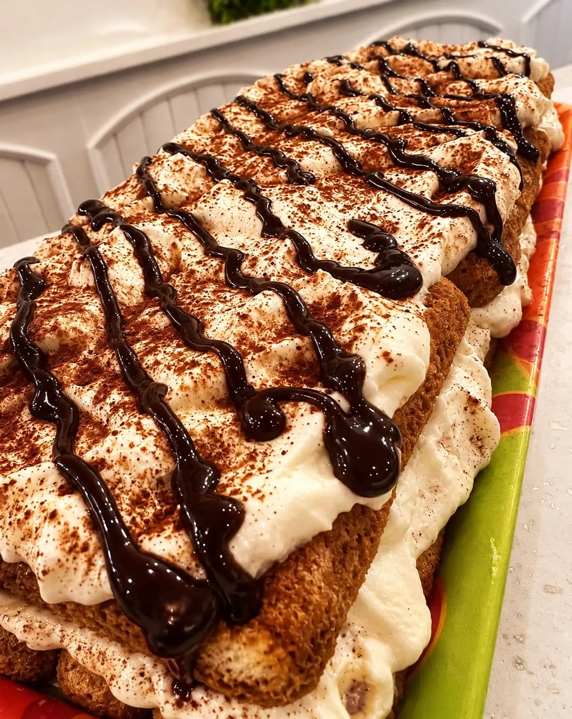 A whole layered sponge and cream cake, dusted with cocoa and drizzled with chocolate, on a long tray.