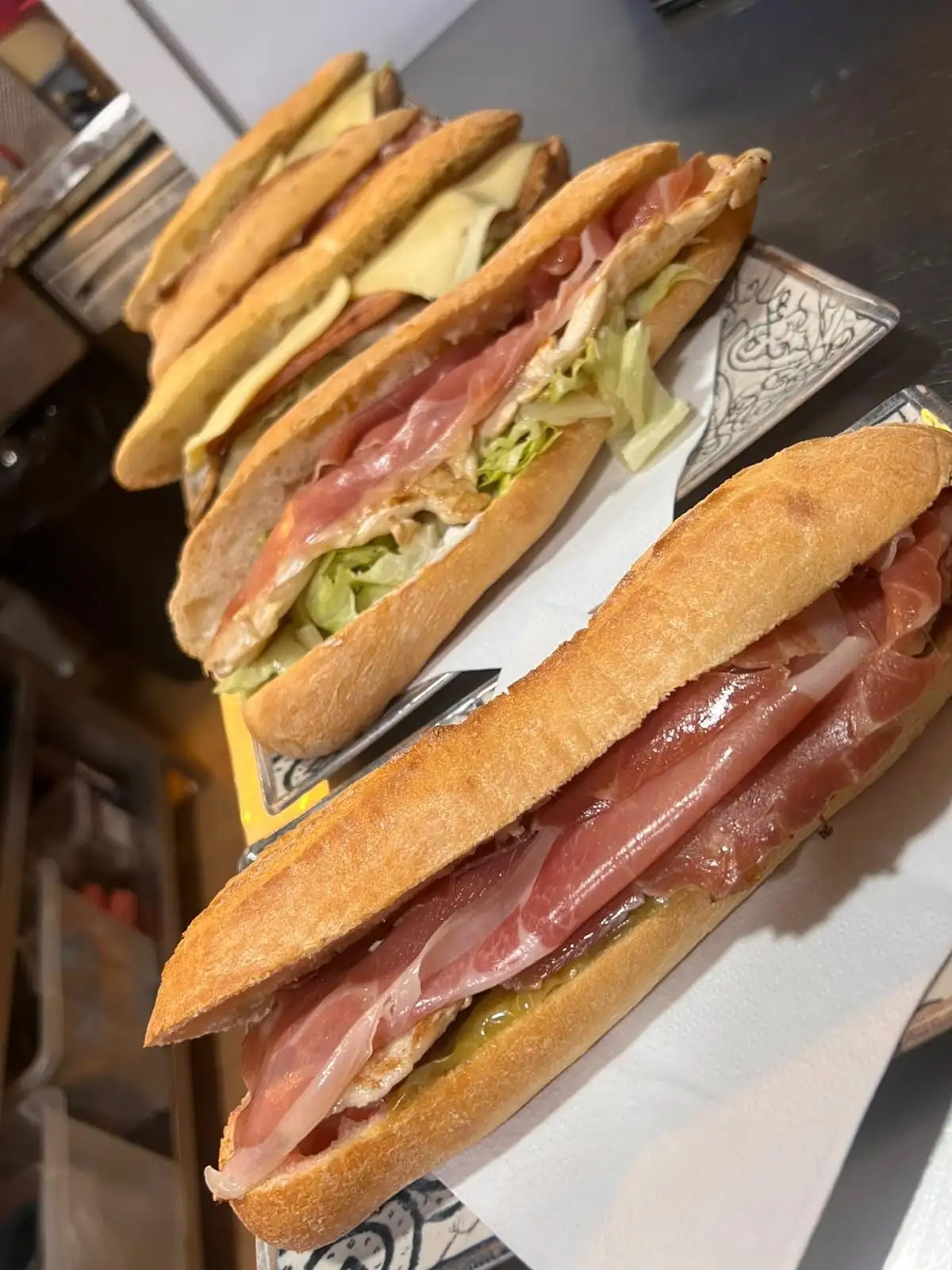 Three baguette sandwiches on napkins: one with serrano ham and egg, one with lettuce and grilled meat, and one with melted cheese.