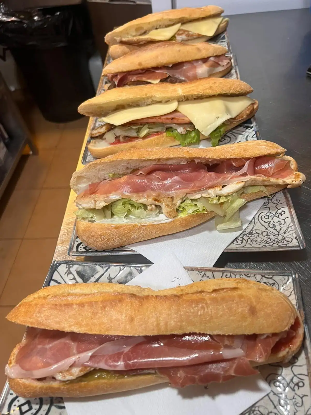 Five baguette sandwiches lined up on trays: serrano ham, grilled meat with lettuce, and melted cheese.