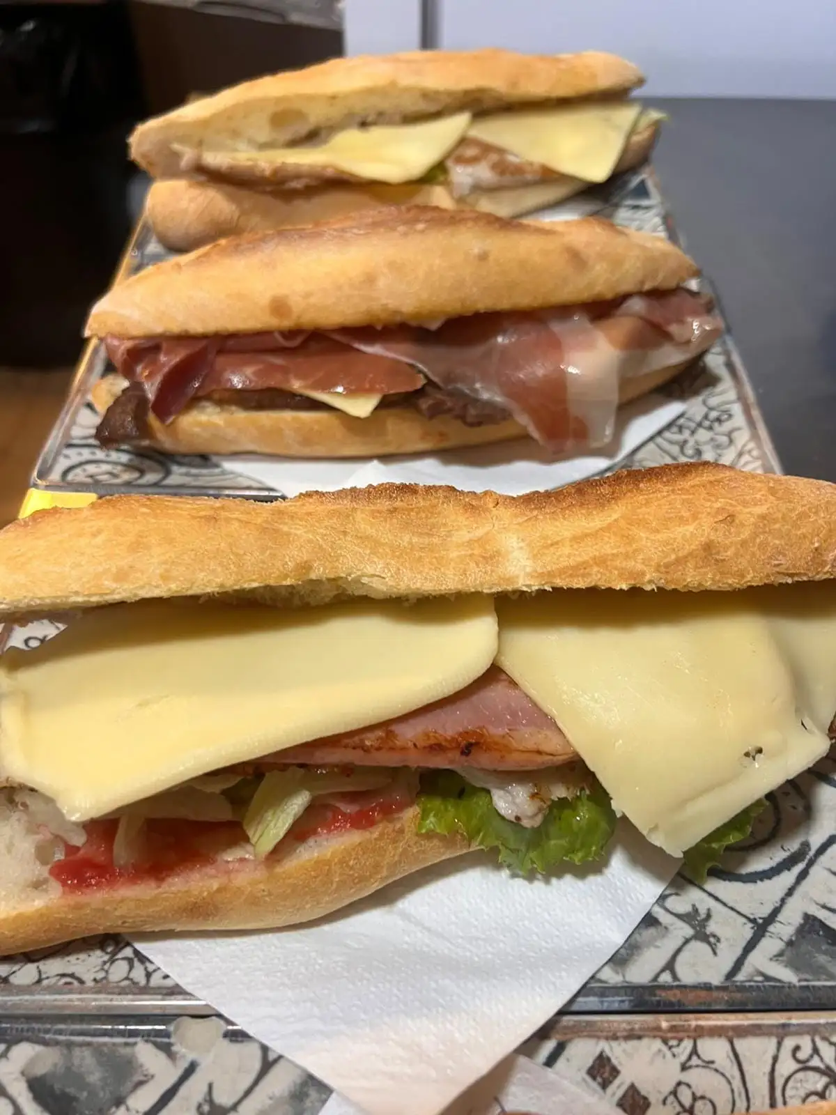 A baguette sandwich with slices of cheese, grilled meat, lettuce and tomato, with two more behind it on the counter.