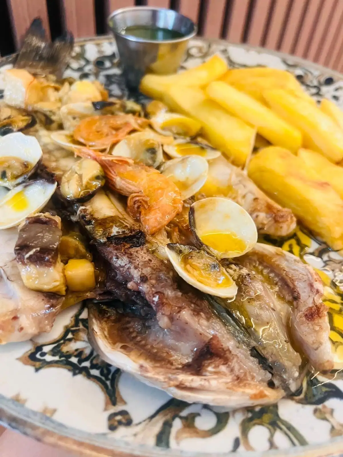 A butterflied grilled fish with prawns, clams and mussels, chips and a small bowl of green sauce, on a patterned plate.