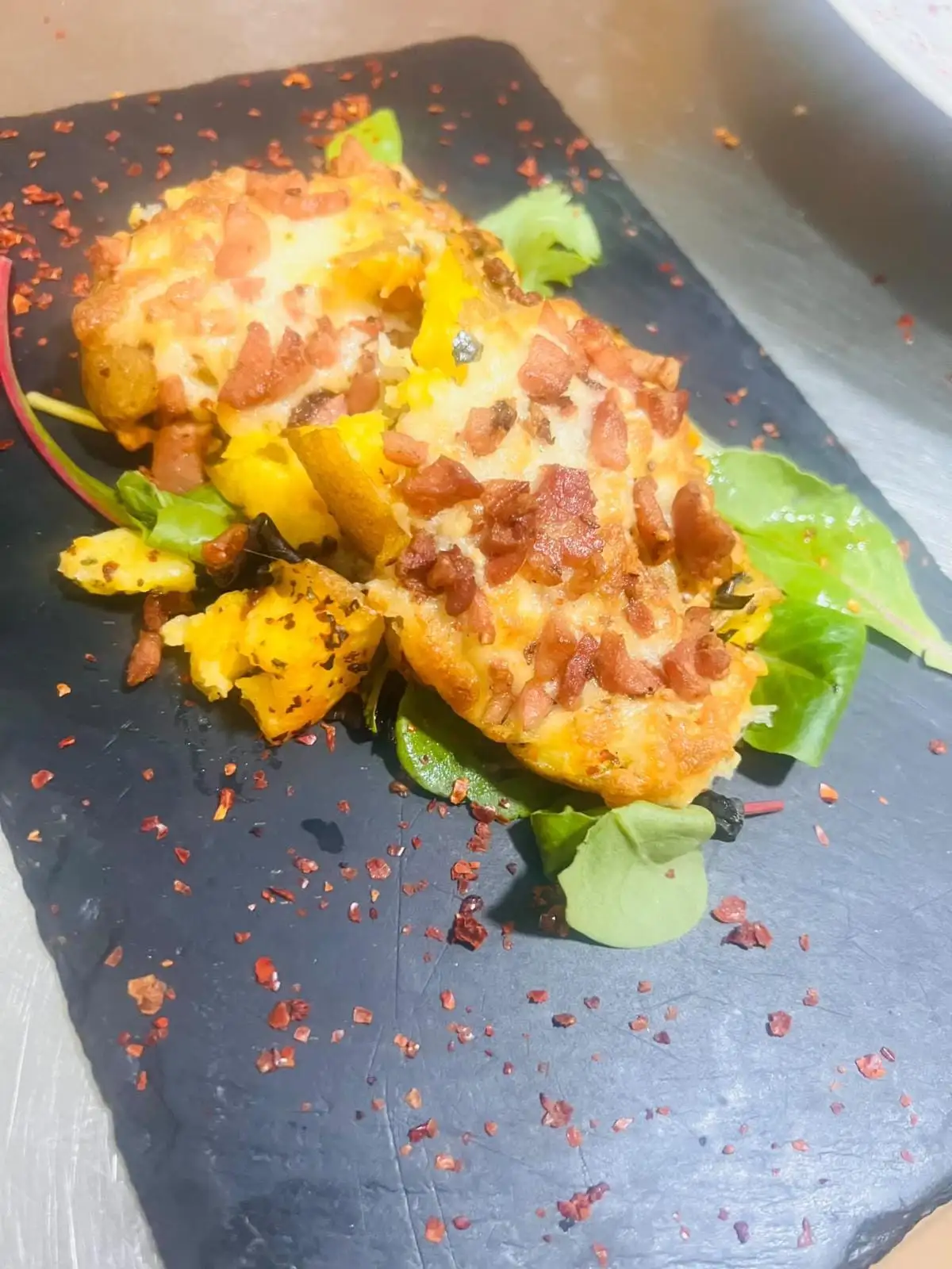 Baked potatoes gratinated with melted cheese and diced bacon, on a slate board with green leaves and red pepper flakes.