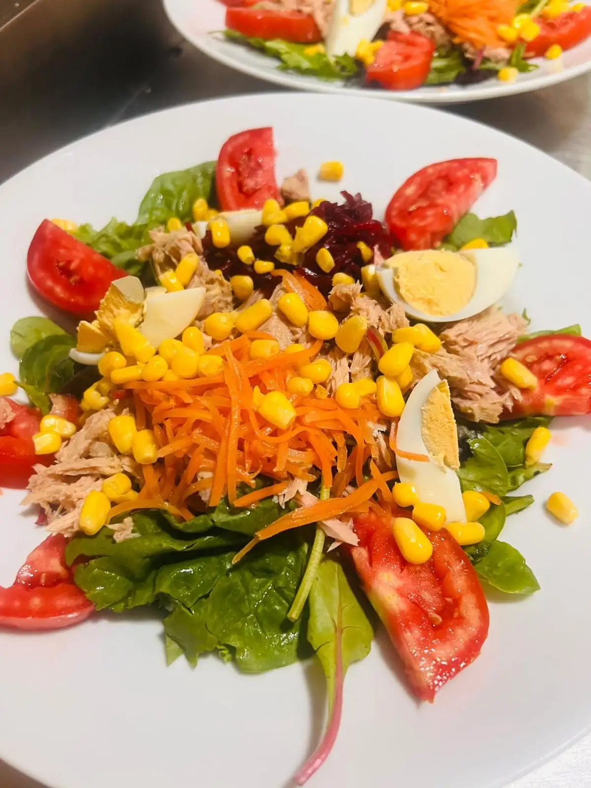 A mixed salad with lettuce, tomato wedges, grated carrot and beetroot, sweetcorn, hard-boiled egg and tuna, with another identical plate behind.