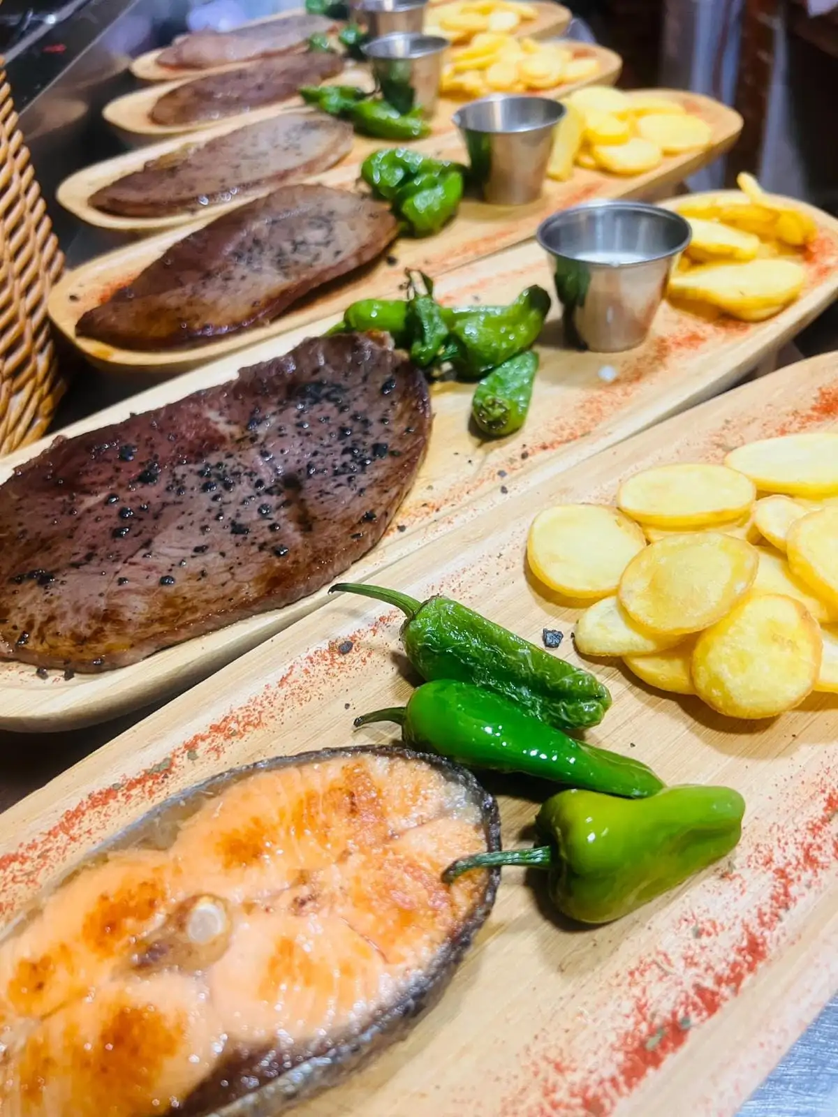 A row of wooden boards with a grilled salmon steak, beef fillets, Padrón peppers, sliced potatoes and little metal sauce cups.