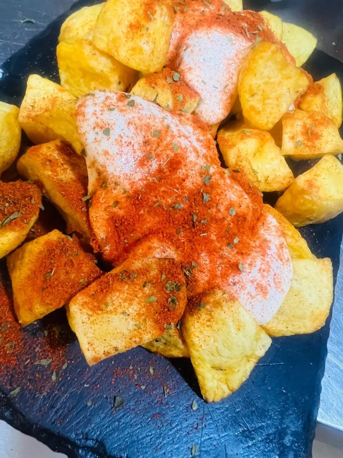 Chunks of fried potato on a slate, topped with white sauce, paprika and oregano.