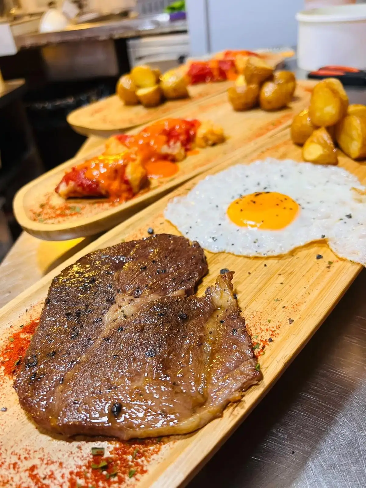 A wooden board with a grilled steak and a fried egg, and more boards behind with roast potatoes and red sauce.