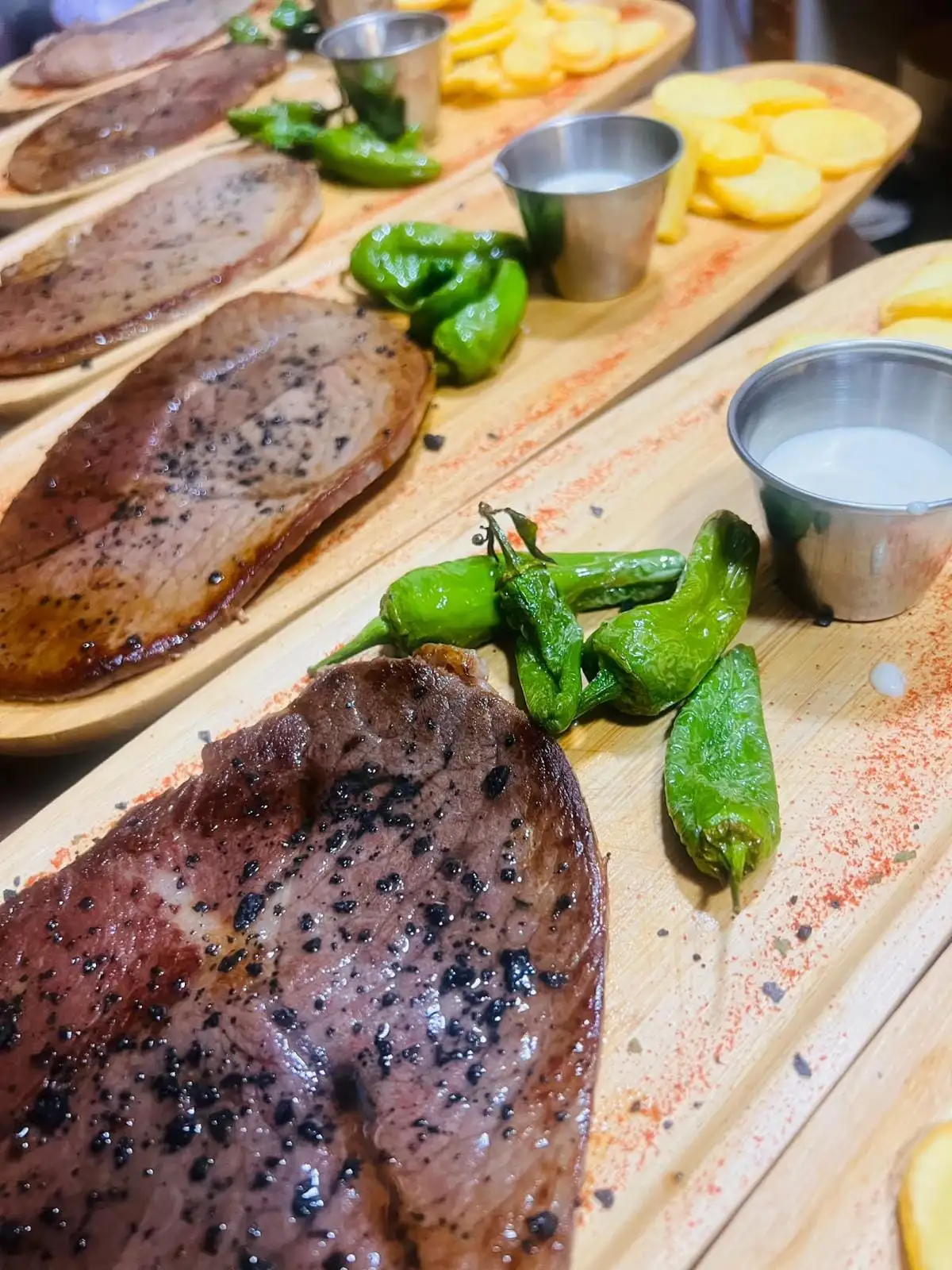 A row of boards with grilled beef, Padrón peppers, small cups of white sauce and sliced potatoes.