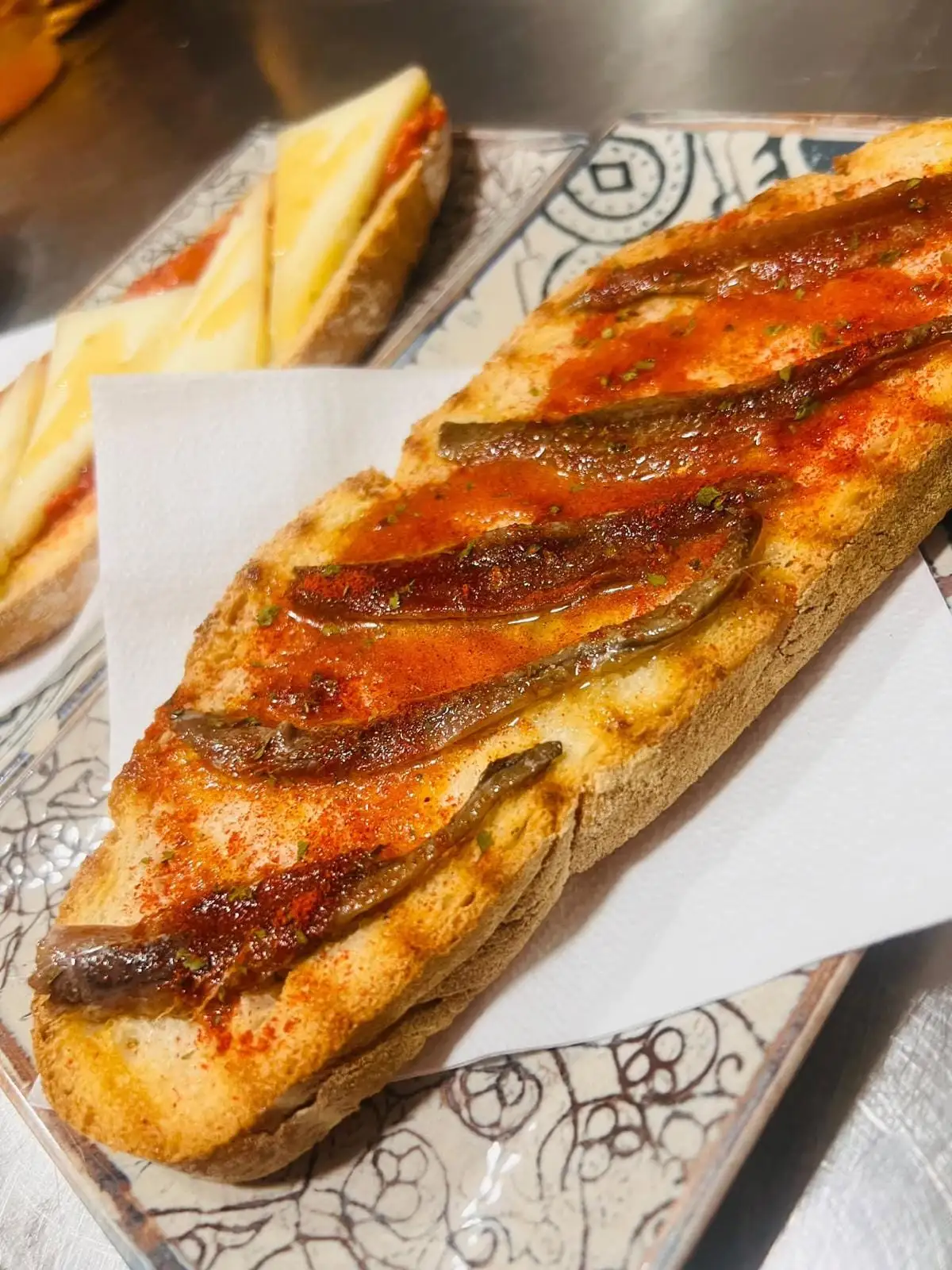 A halved baguette, toasted, with tomato, anchovies and paprika, and behind it another with tomato and sliced cheese.