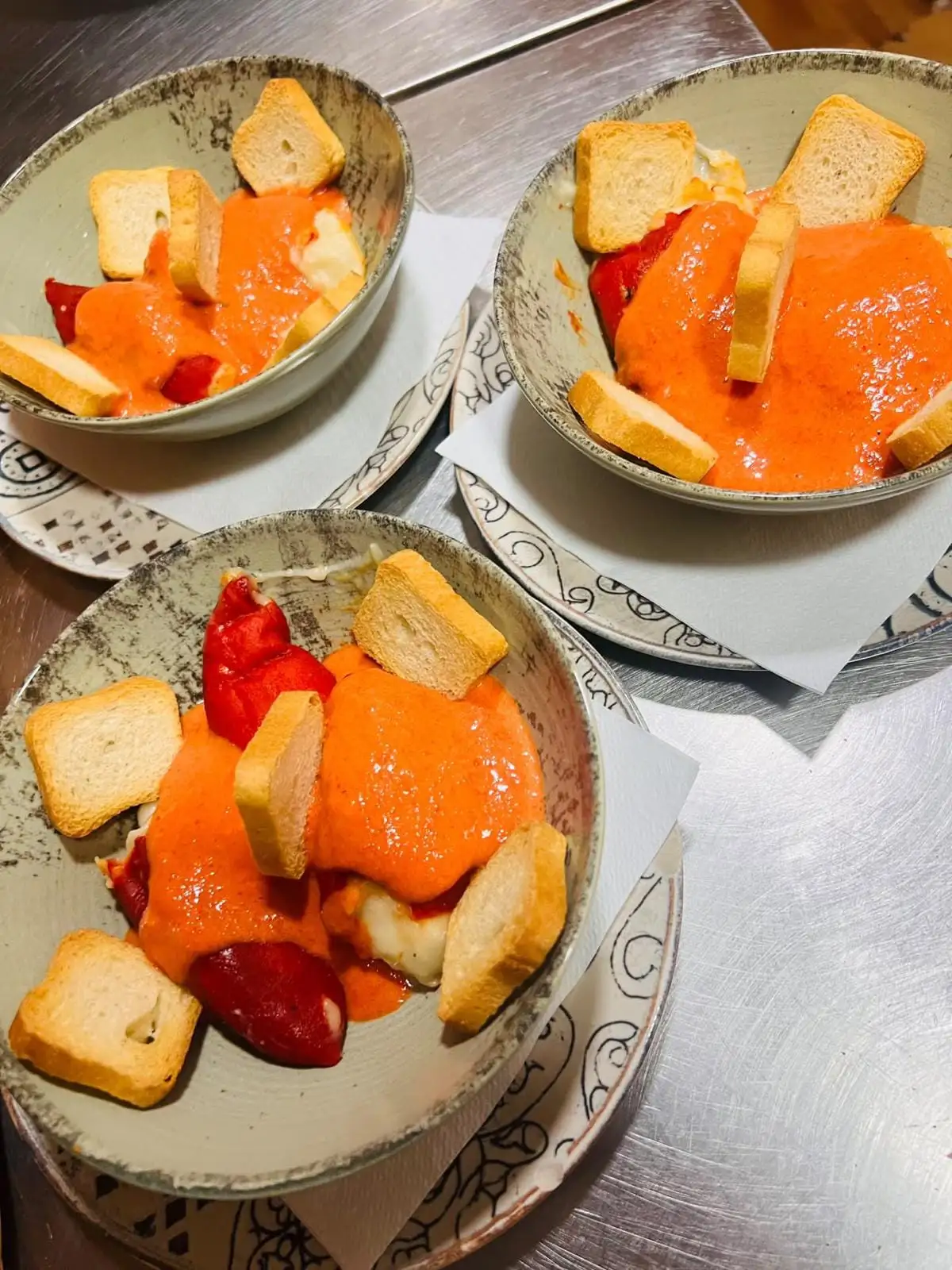 Three bowls of roasted red peppers and cheese under a creamy tomato sauce, with slices of toasted bread around the edge.