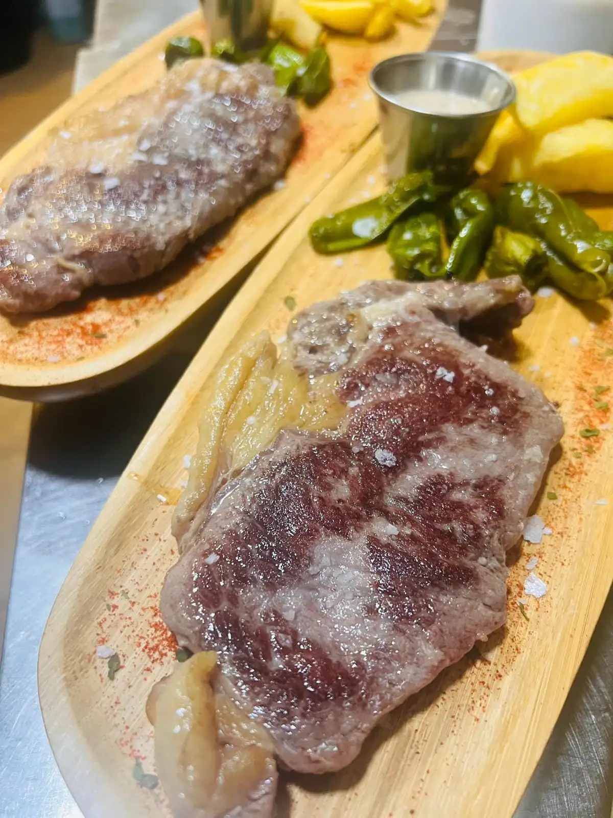 Two grilled rib steaks with coarse salt on wooden boards, with Padrón peppers, chips and a small cup of white sauce.