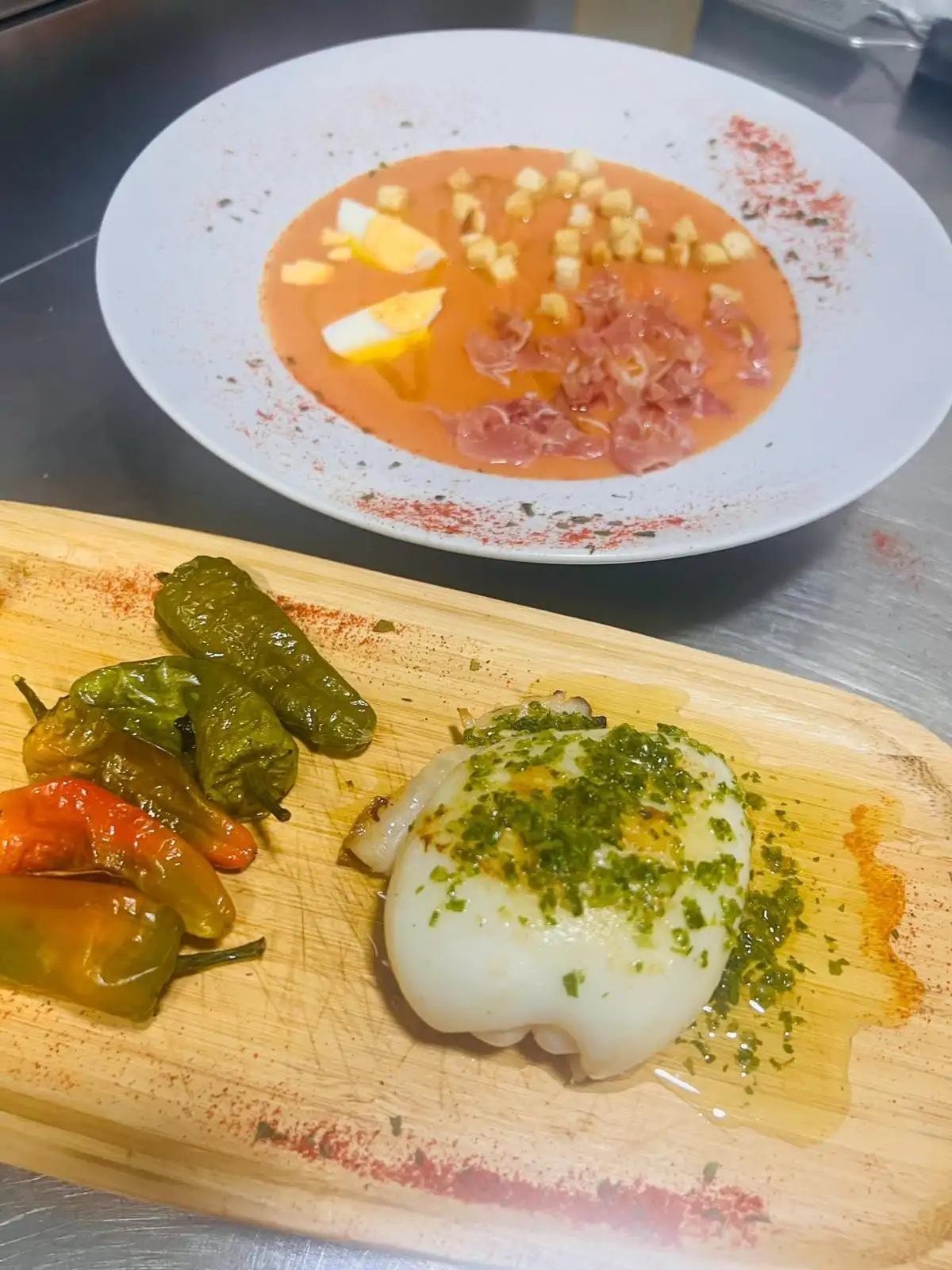 A board with grilled cuttlefish in garlic and parsley and roasted peppers, and behind it a plate of chilled tomato soup with hard-boiled egg, croutons and ham.