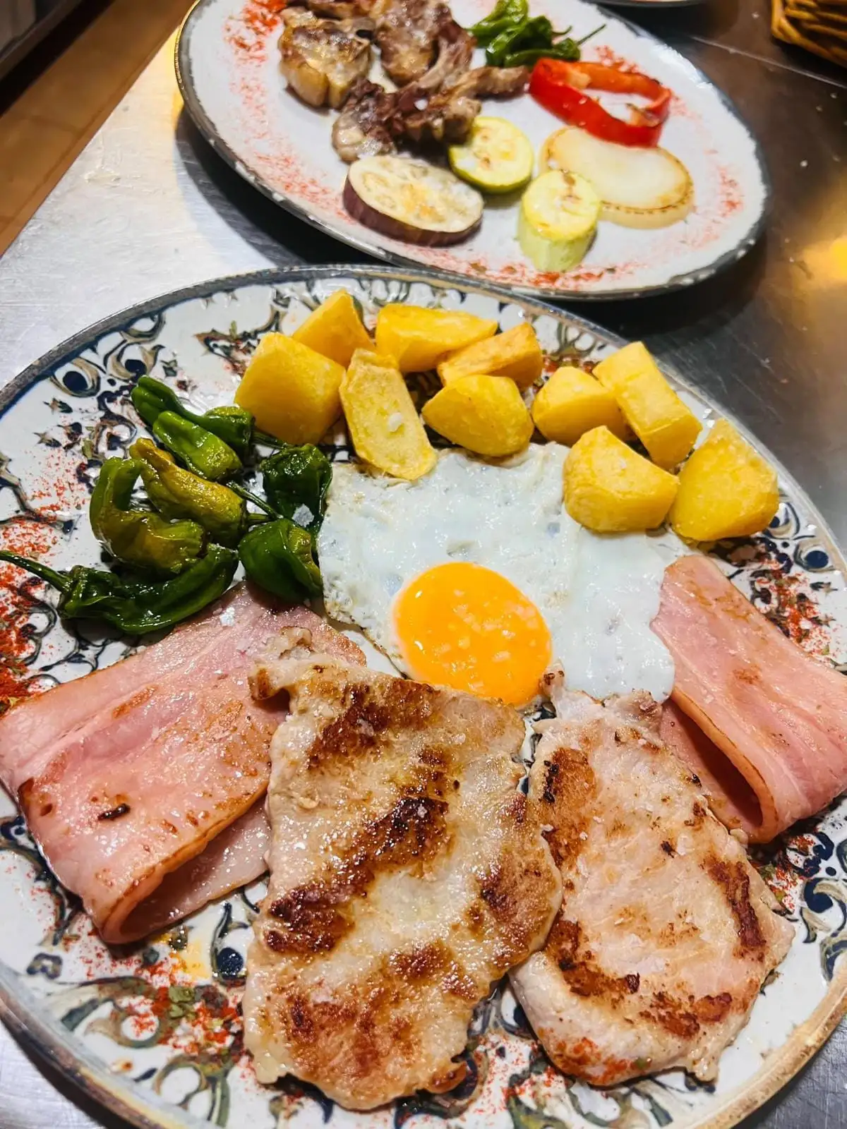 A plate with grilled fillets, slices of cooked ham, a fried egg, chunky chips and Padrón peppers, and another plate of grilled meat and vegetables behind.