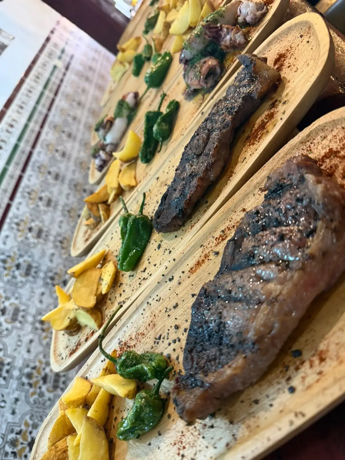 A row of boards with grilled steaks, Padrón peppers, potato wedges and, further back, squid with green sauce.