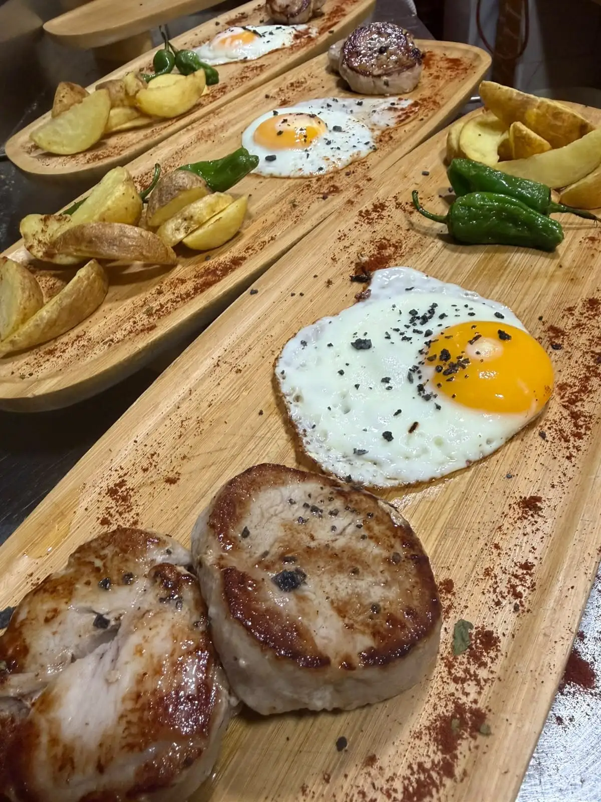 Wooden boards lined up, each with two round cuts of grilled meat, a fried egg, potato wedges and green peppers.