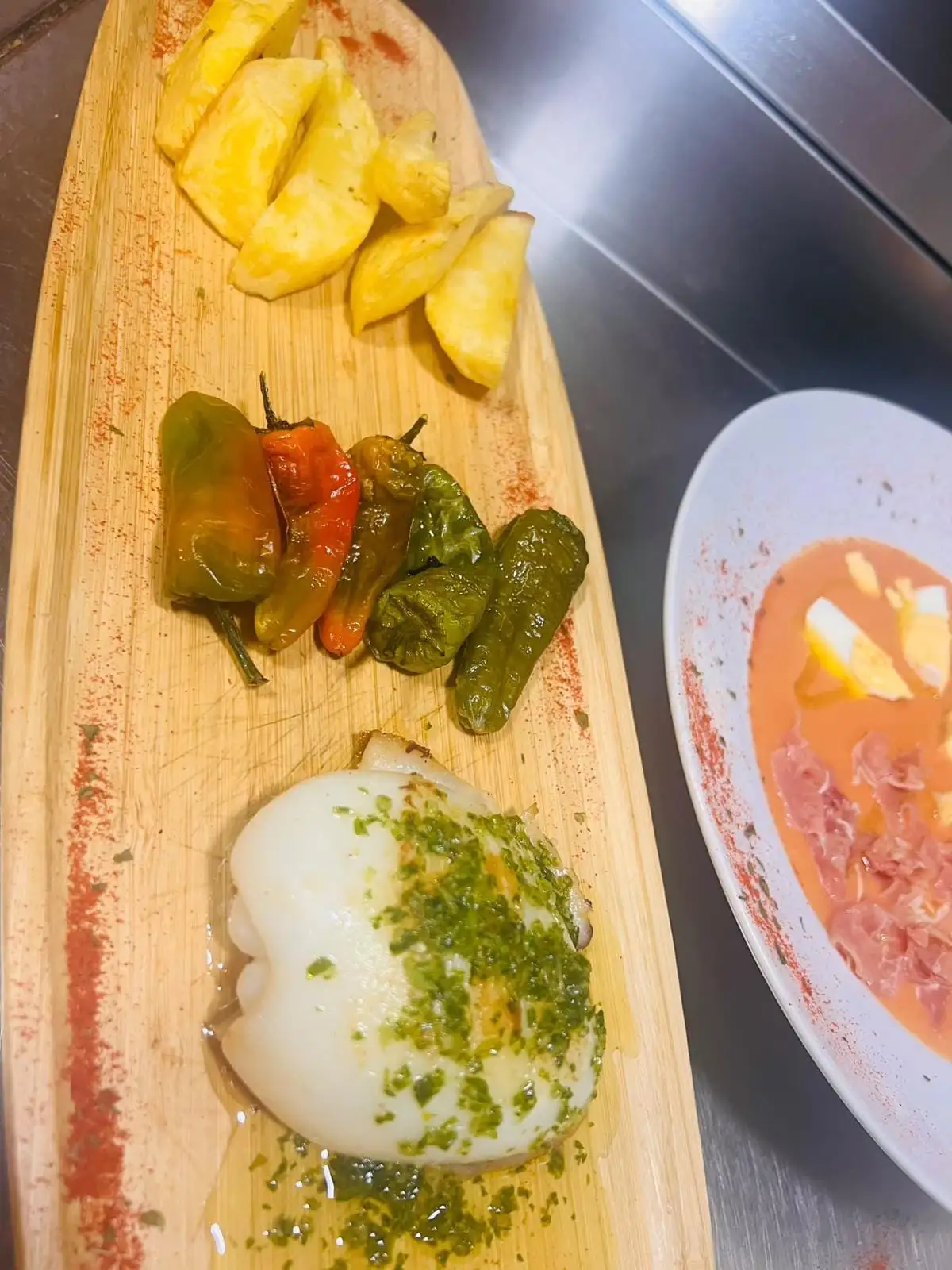 A wooden board with grilled cuttlefish in green sauce, Padrón peppers and fried potatoes, next to a plate of salmorejo with hard-boiled egg and ham.