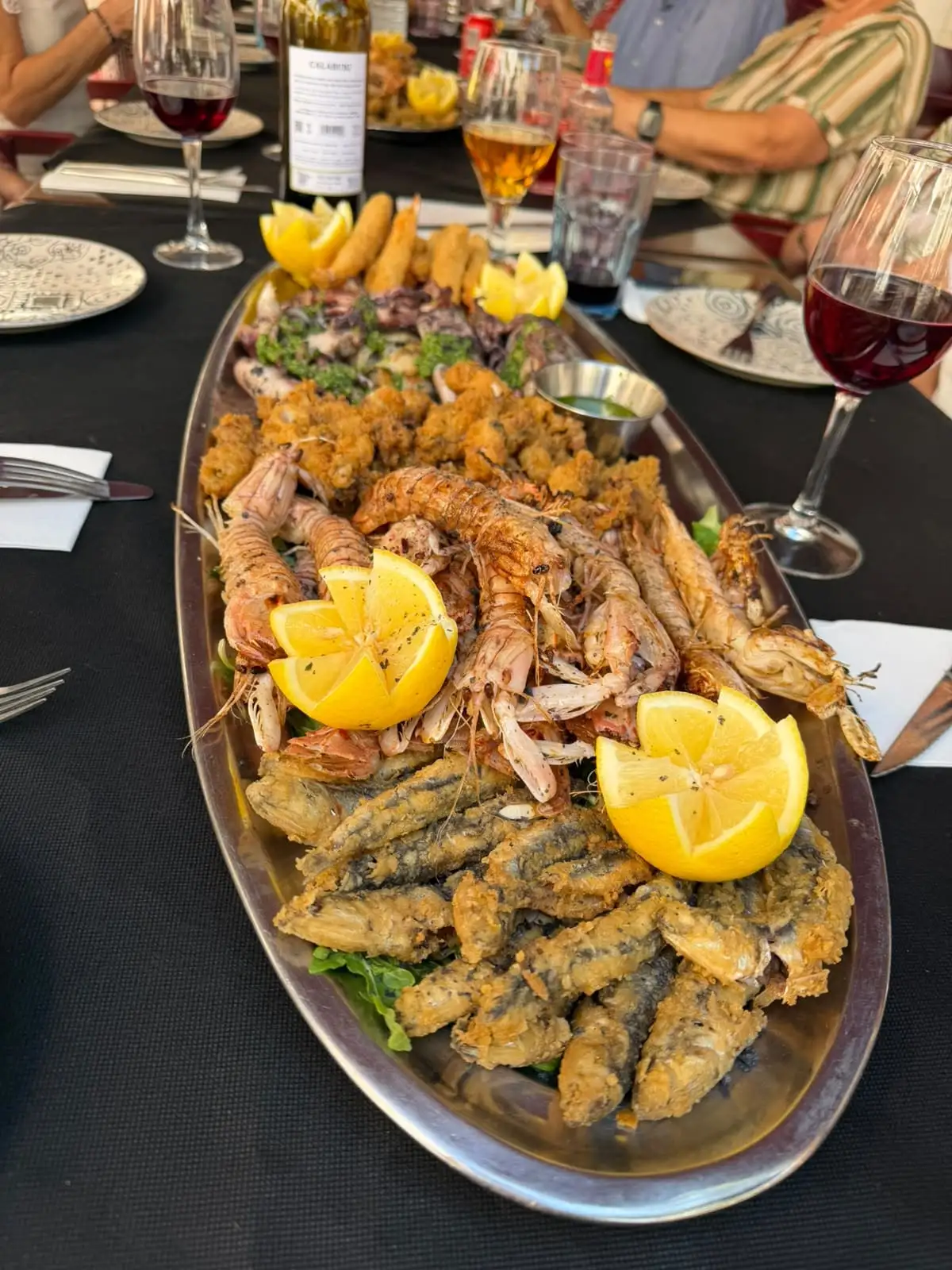 A large oval seafood platter in the middle of the table: grilled mantis shrimp and prawns, fried anchovies and squid, lemon and a small bowl of green sauce.