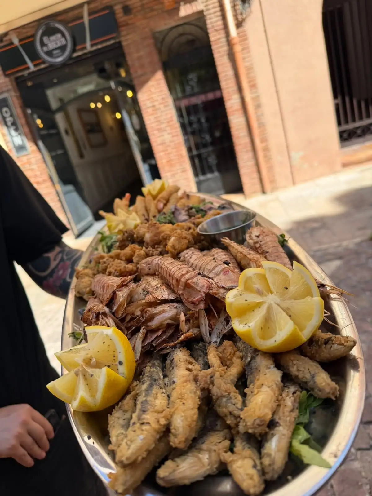 A seafood platter held out in the street, with the sign and front of the bar behind: mantis shrimp, prawns, fried anchovies and squid with lemon.