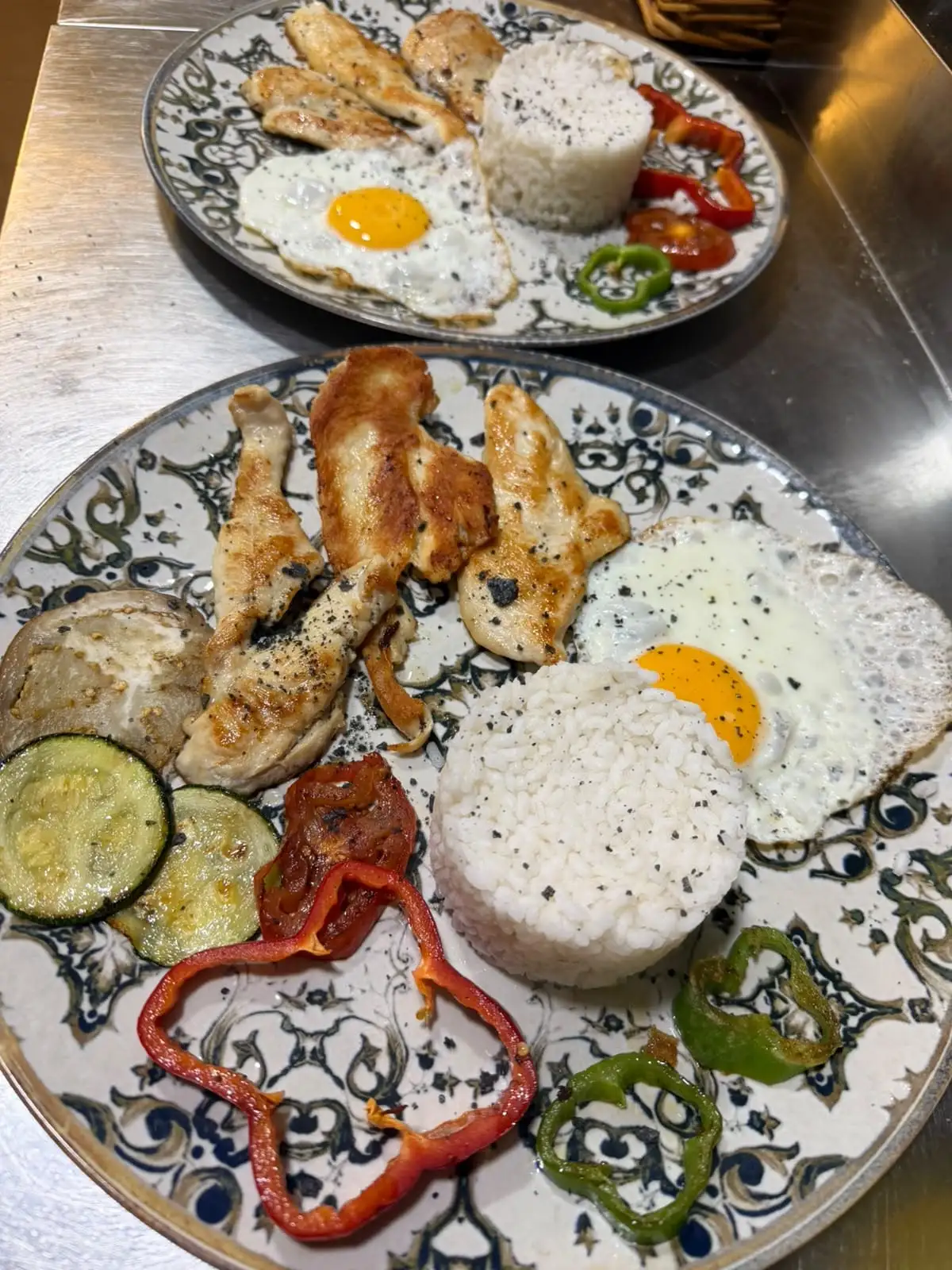 Two patterned plates with strips of grilled meat, a fried egg, a mound of white rice and roasted vegetables.