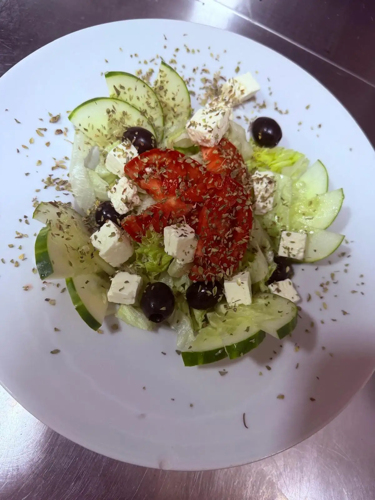 A salad on a white plate: lettuce, cucumber slices, cubes of feta, black olives and tomato, sprinkled with oregano.