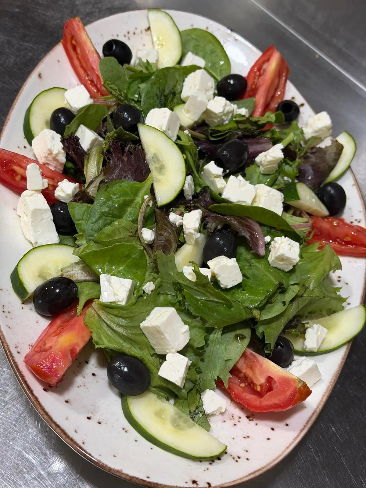 A mixed-leaf salad on an oval plate, with cubes of feta, black olives, tomato wedges and cucumber slices.