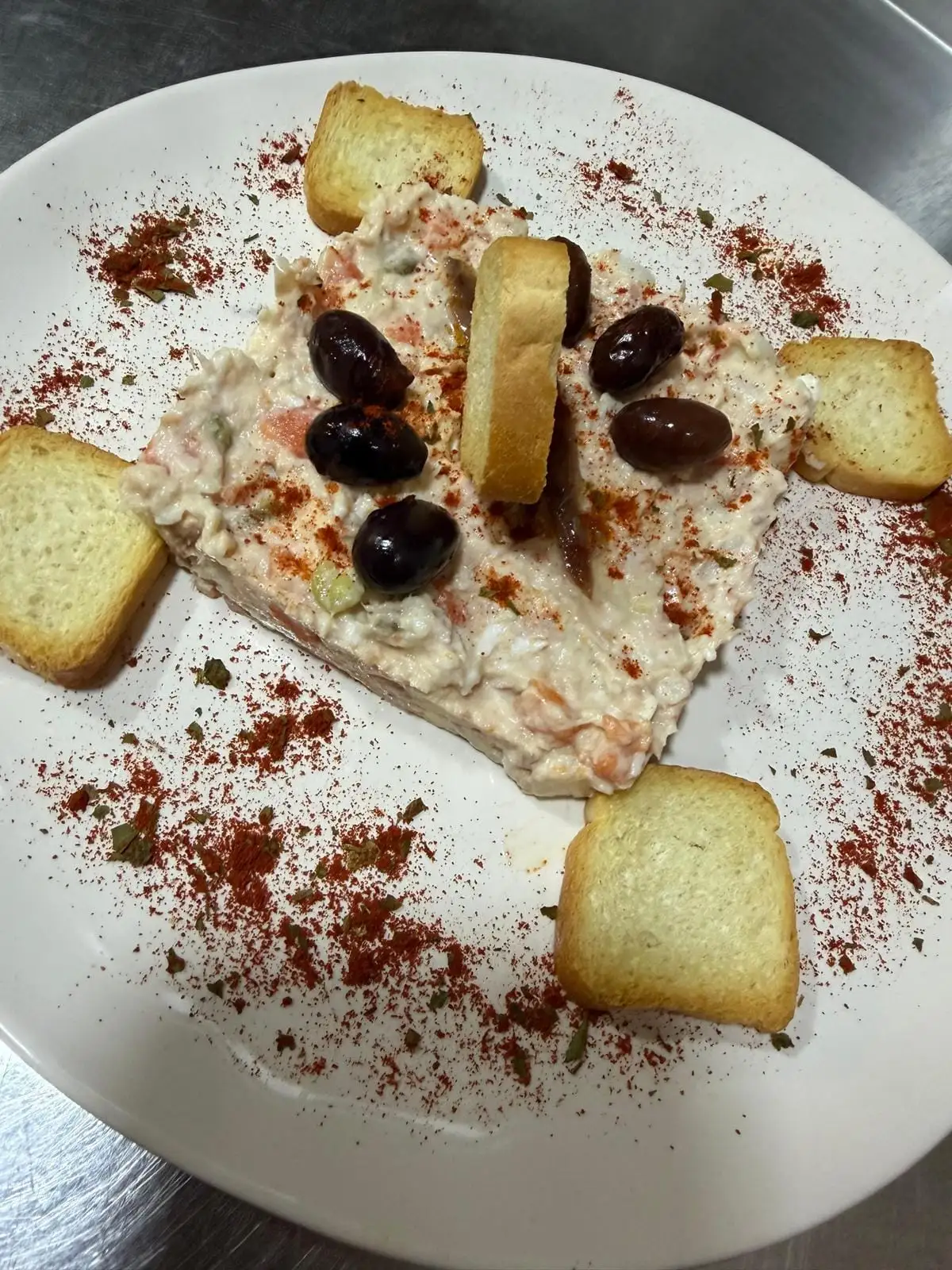A moulded block of creamy salad in the middle of the plate, topped with black olives and anchovies, toasts around it and a dusting of paprika.