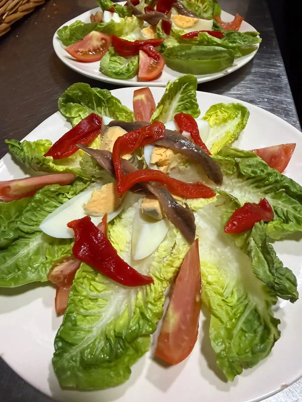 Two plates of salad with baby gem leaves, tomato wedges, hard-boiled egg, strips of roasted red pepper and anchovies.