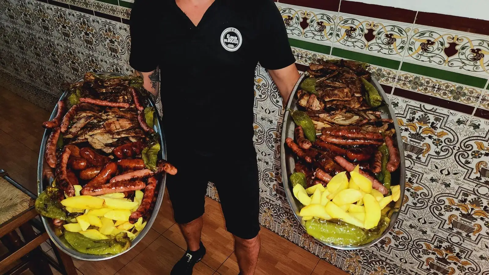 Two large oval platters of mixed grill held up in the dining room, with sausage, chorizo, grilled meat, chips and green peppers.