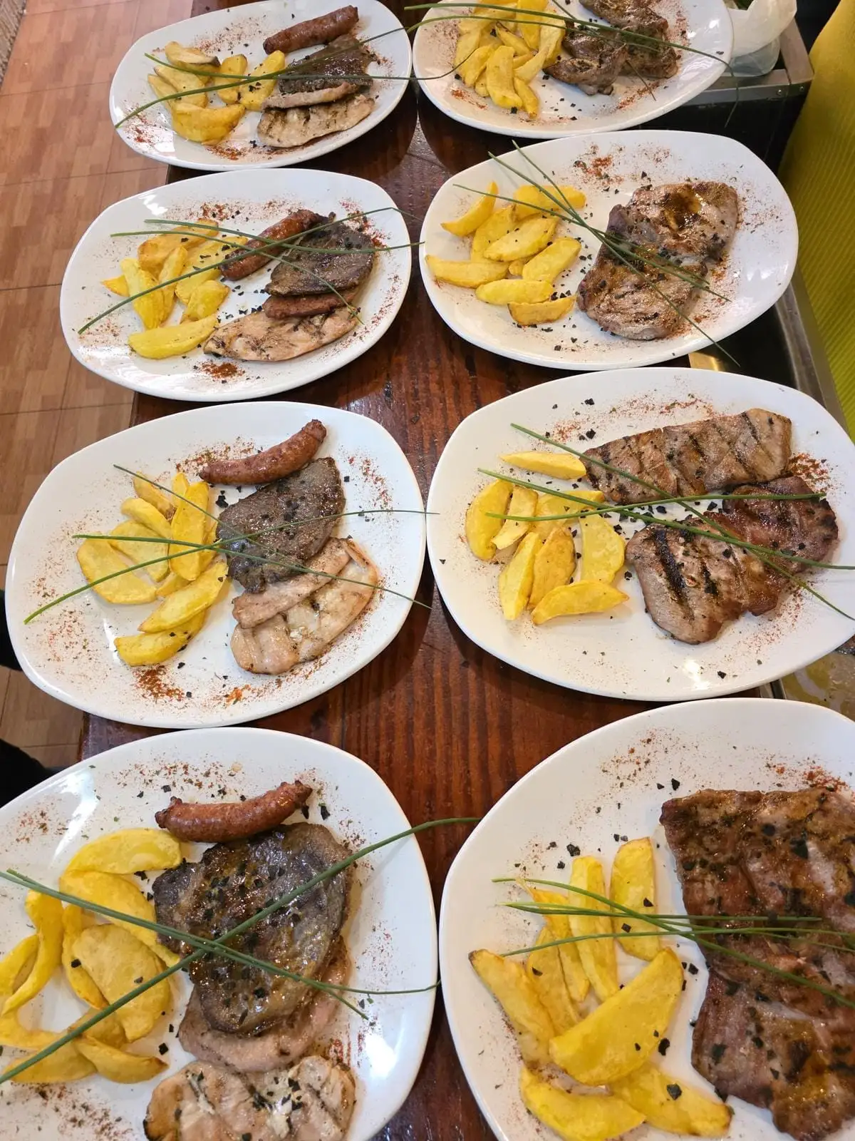Several plates lined up on a wooden table, each with grilled meat, potato wedges and paprika dusted around the rim.