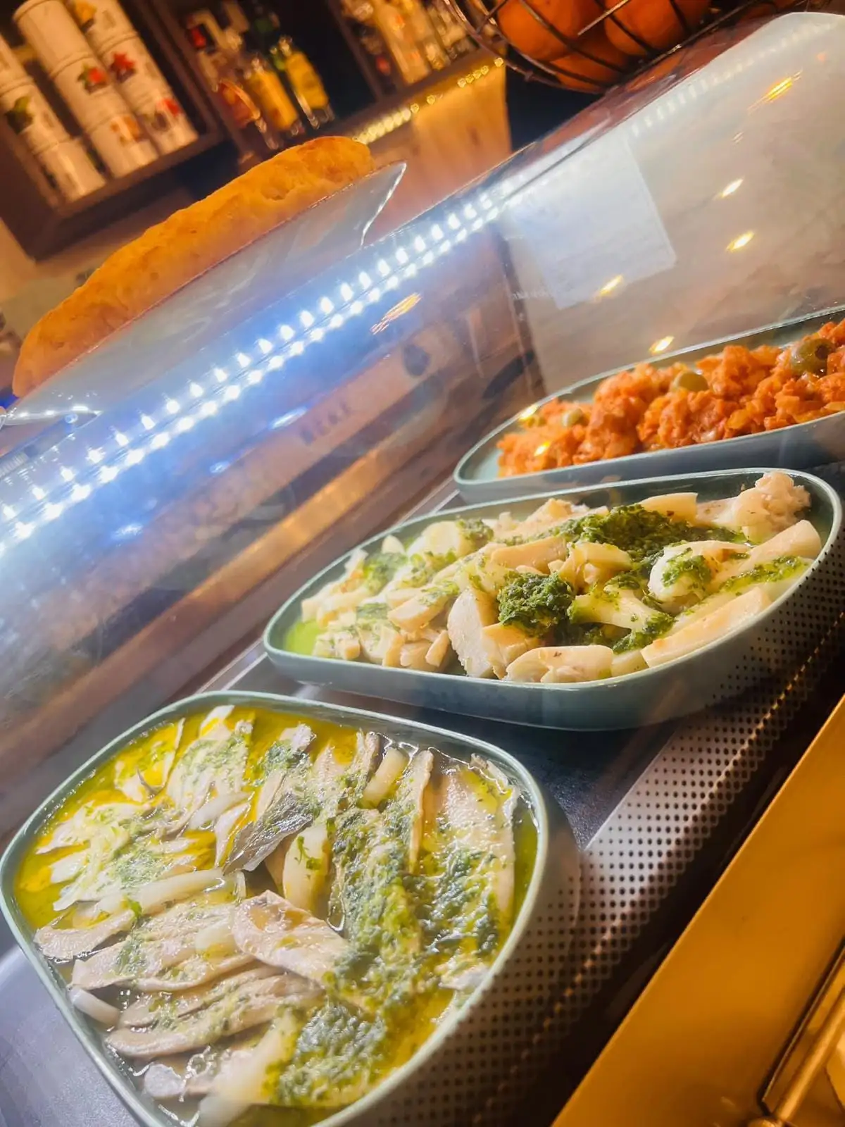Tapas display case at the bar, with trays of fish in oil and parsley, another in green sauce and another with tomato and olives, and a loaf resting on the glass.