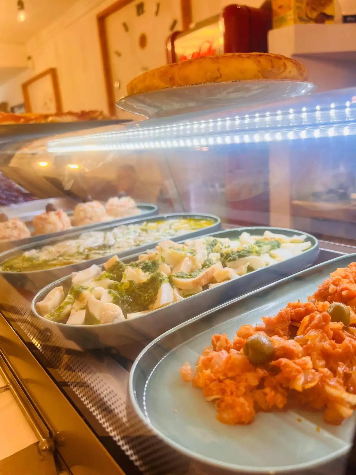 Trays of tapas lined up inside the display case: fish in oil, pieces in green sauce, a tomato and olive salad and, behind them, portions topped with a prawn.
