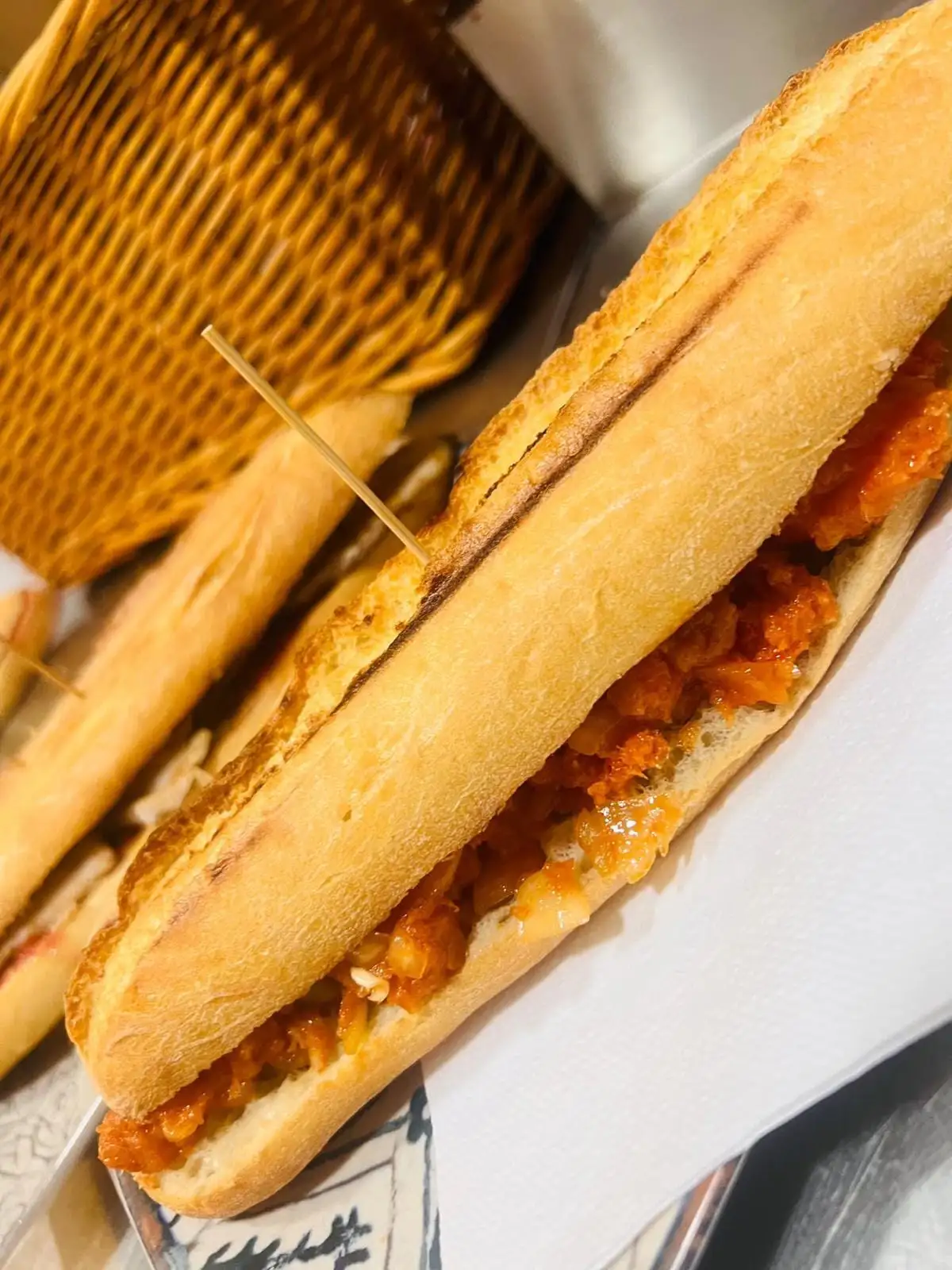Baguette rolls with a filling in tomato sauce, held together with cocktail sticks and served on paper beside a wicker basket.