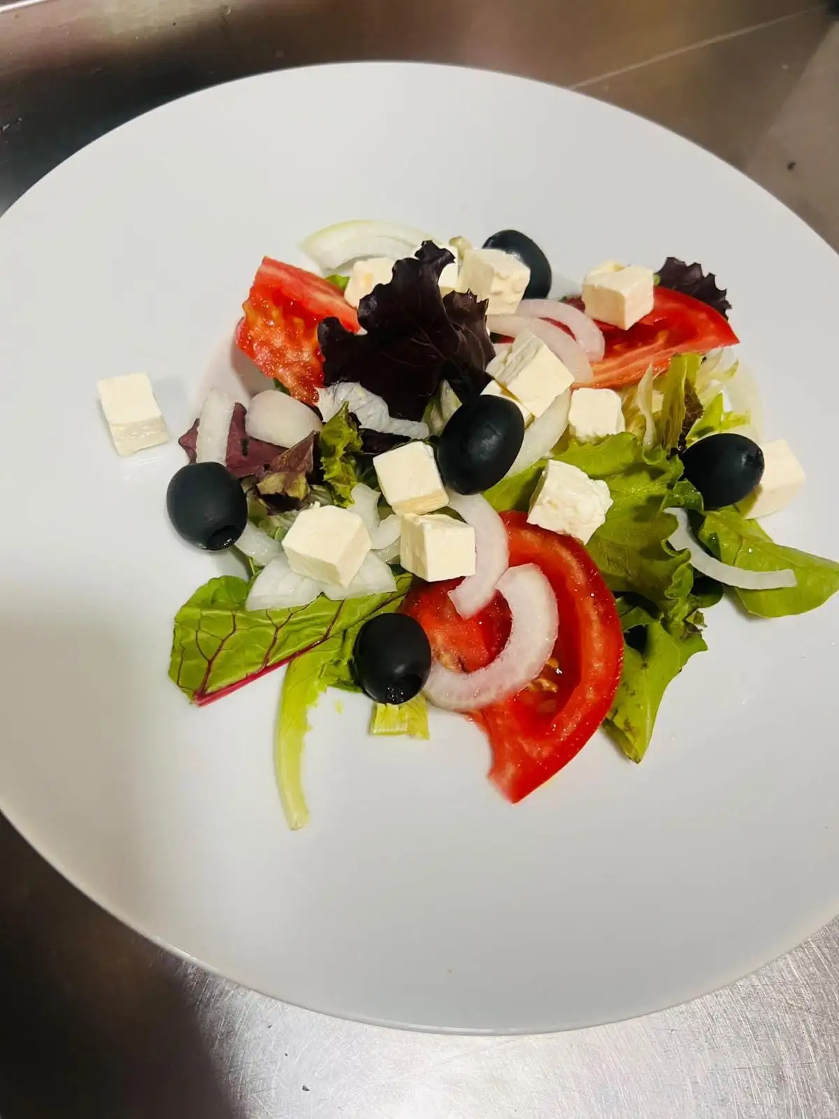 A mixed leaf salad with tomato wedges, onion rings, black olives and cubes of white cheese, on a white plate.