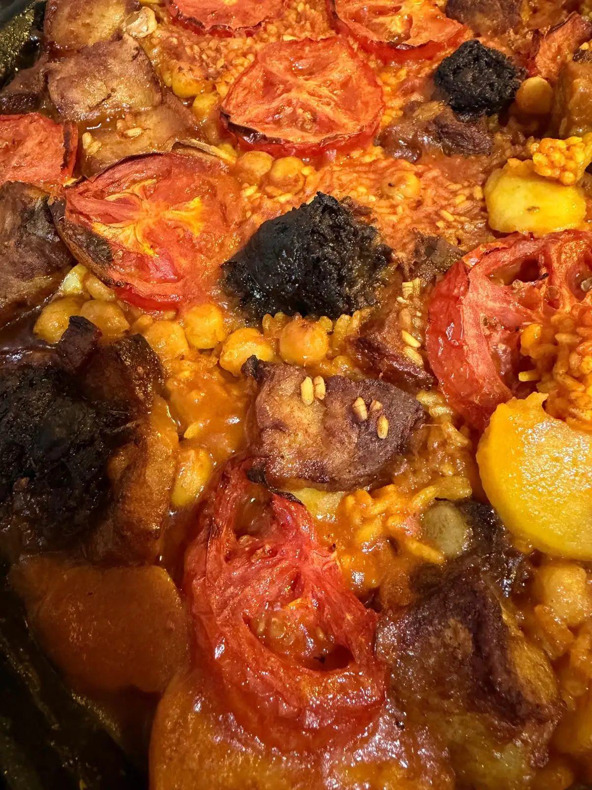 Close-up of a baked rice in its dish: slices of roasted tomato, pieces of blood sausage, chickpeas, chunks of meat and rounds of potato among the toasted grains.