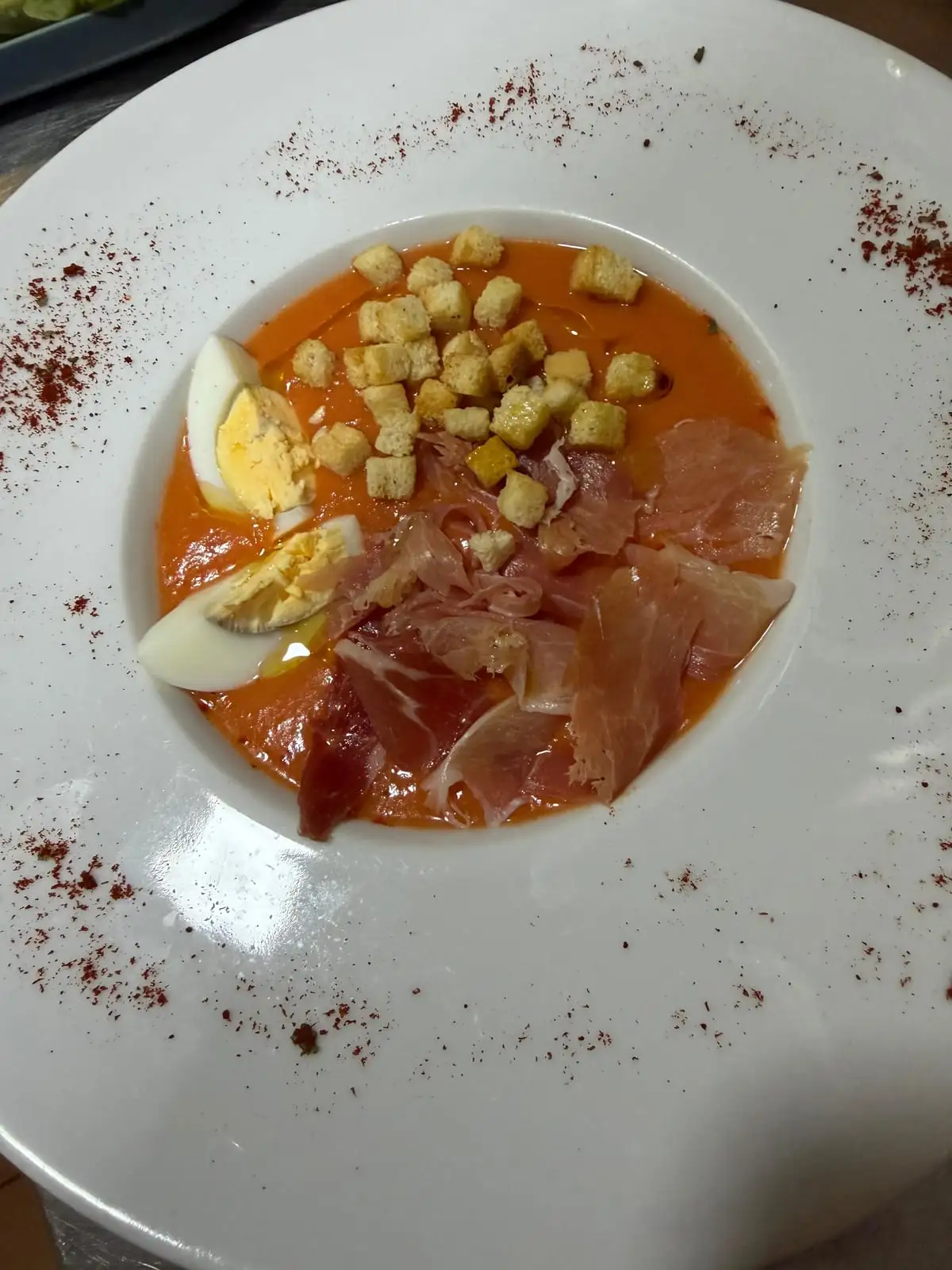 A white soup plate with chilled tomato cream, quarters of hard-boiled egg, croutons and slices of serrano ham, with paprika dusted all around the rim.