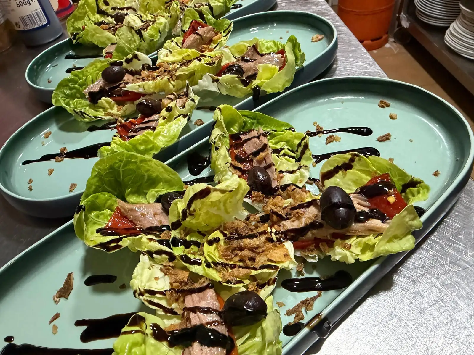 A row of green oval plates with baby gem leaves filled with tuna, tomato and black olive, topped with crisp fried onion and streaks of balsamic reduction.