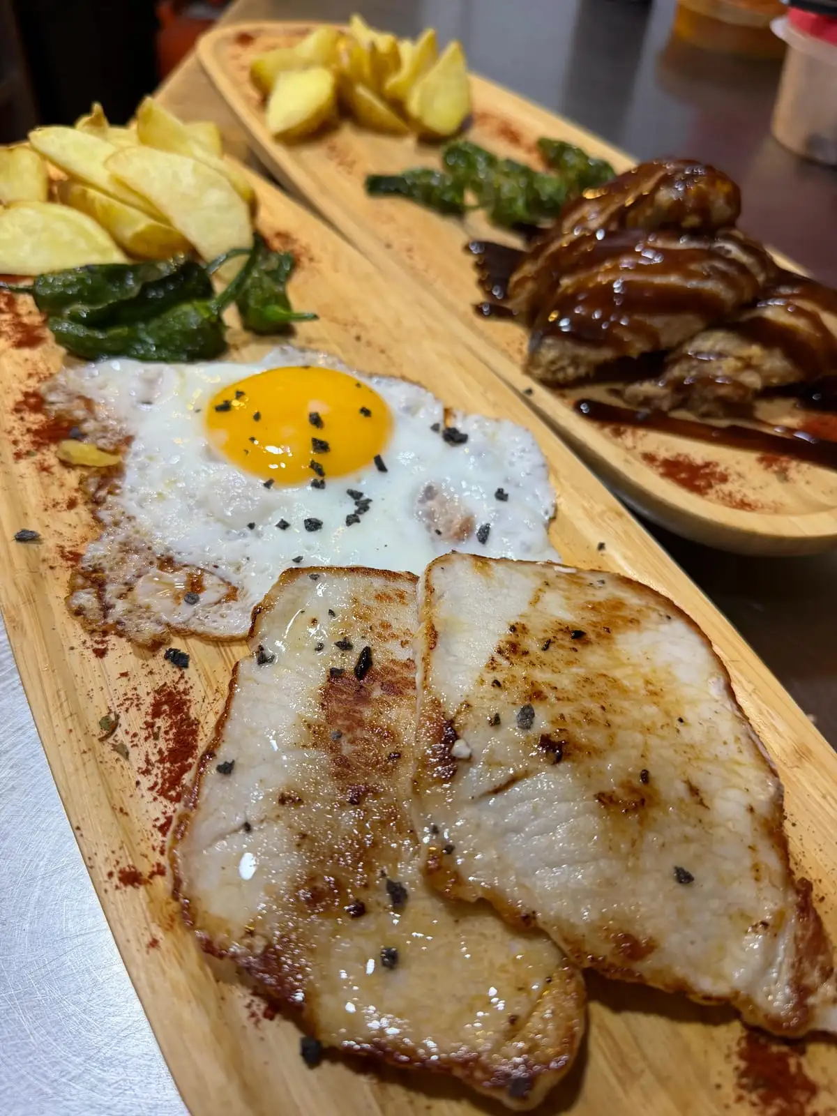 A long wooden board with two grilled pork loin steaks, a fried egg, fried potatoes and Padrón peppers; behind it, another board with meat in a dark sauce and potato wedges.