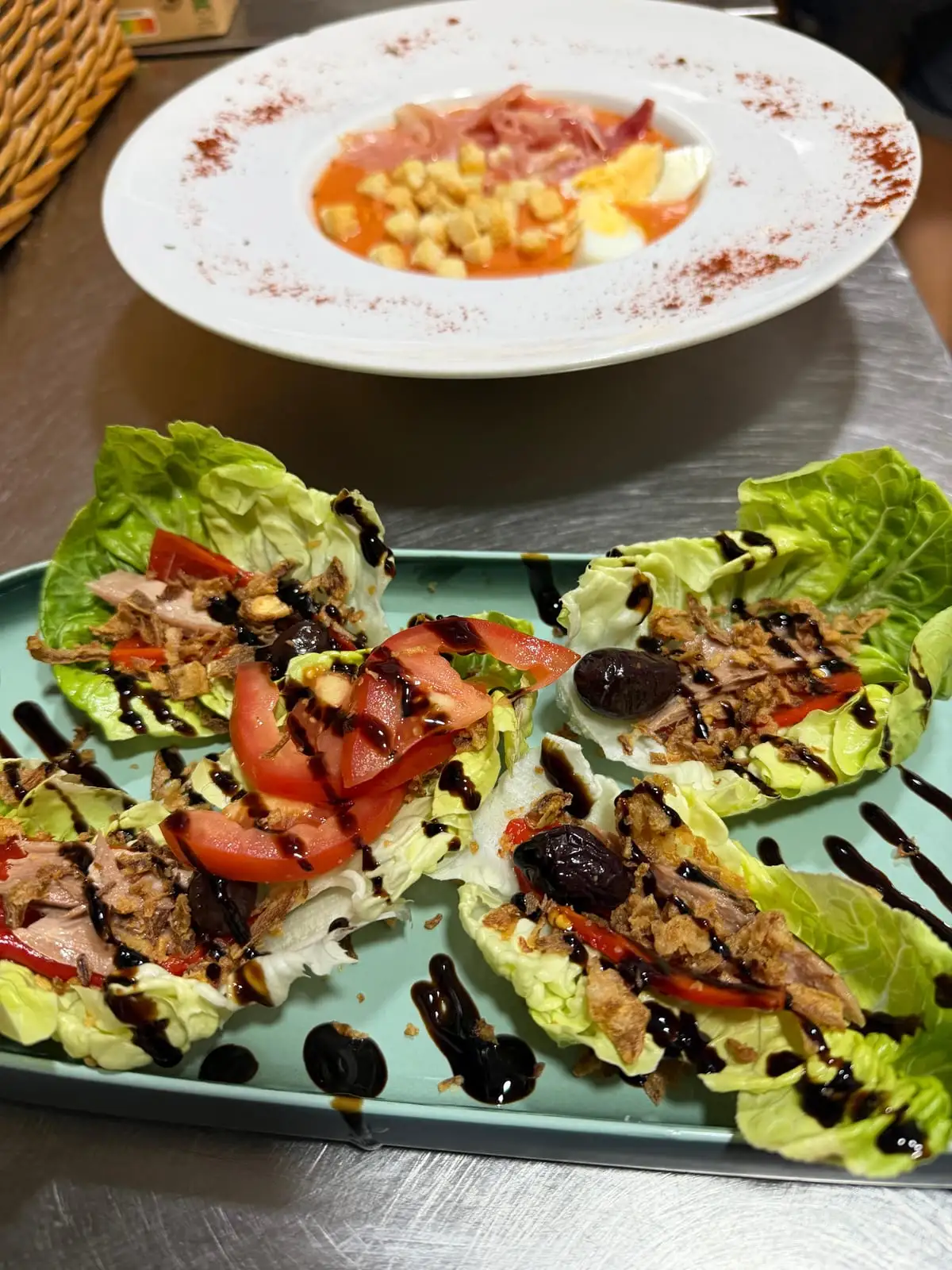 A green tray with four baby gem leaves filled with tuna, tomato and black olive under a balsamic reduction, and behind it a plate of chilled tomato cream with hard-boiled egg, croutons and ham.