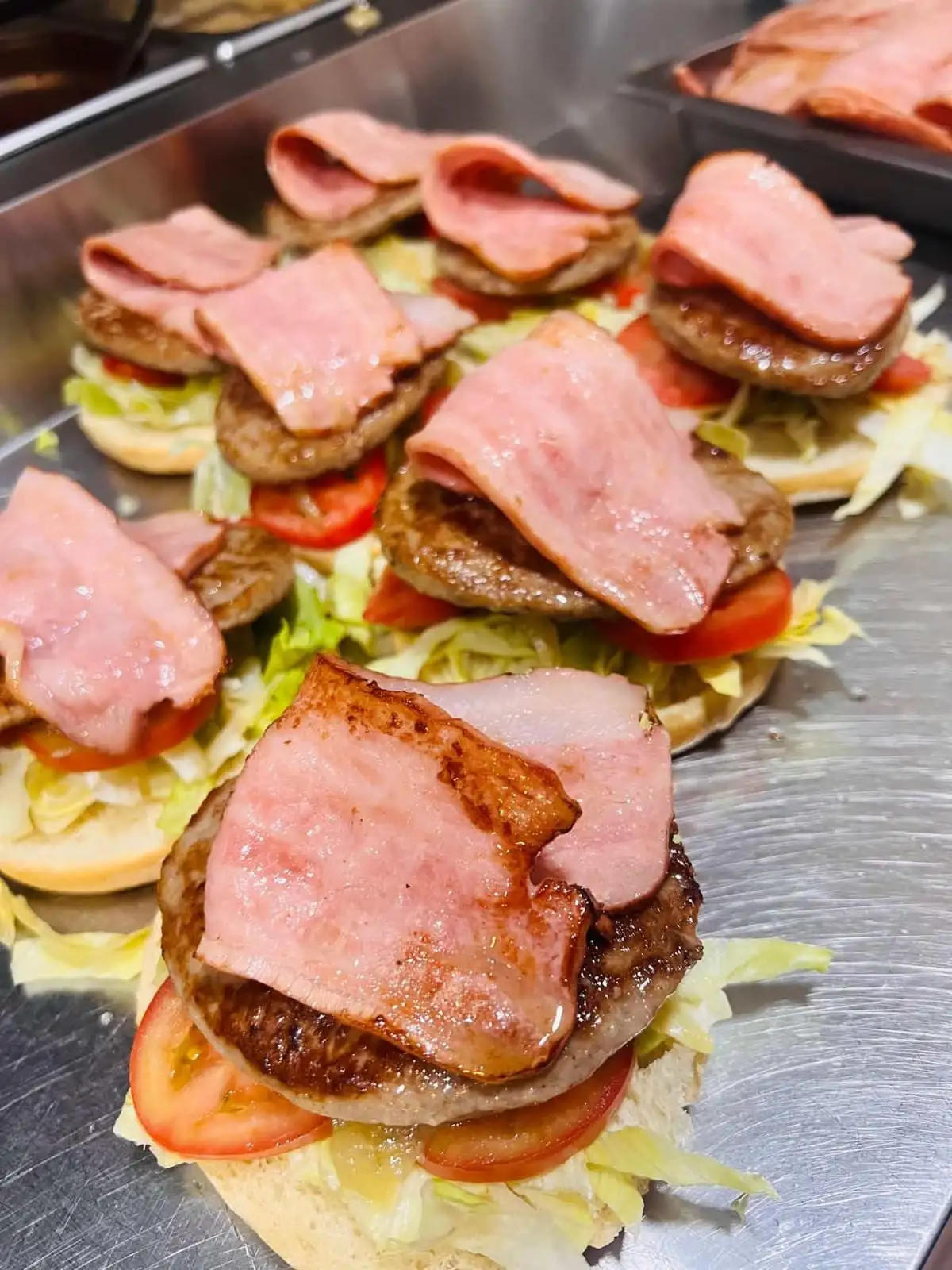 Round buns opened in a row on the griddle, each with lettuce, tomato slices, a grilled burger and rashers of bacon on top.
