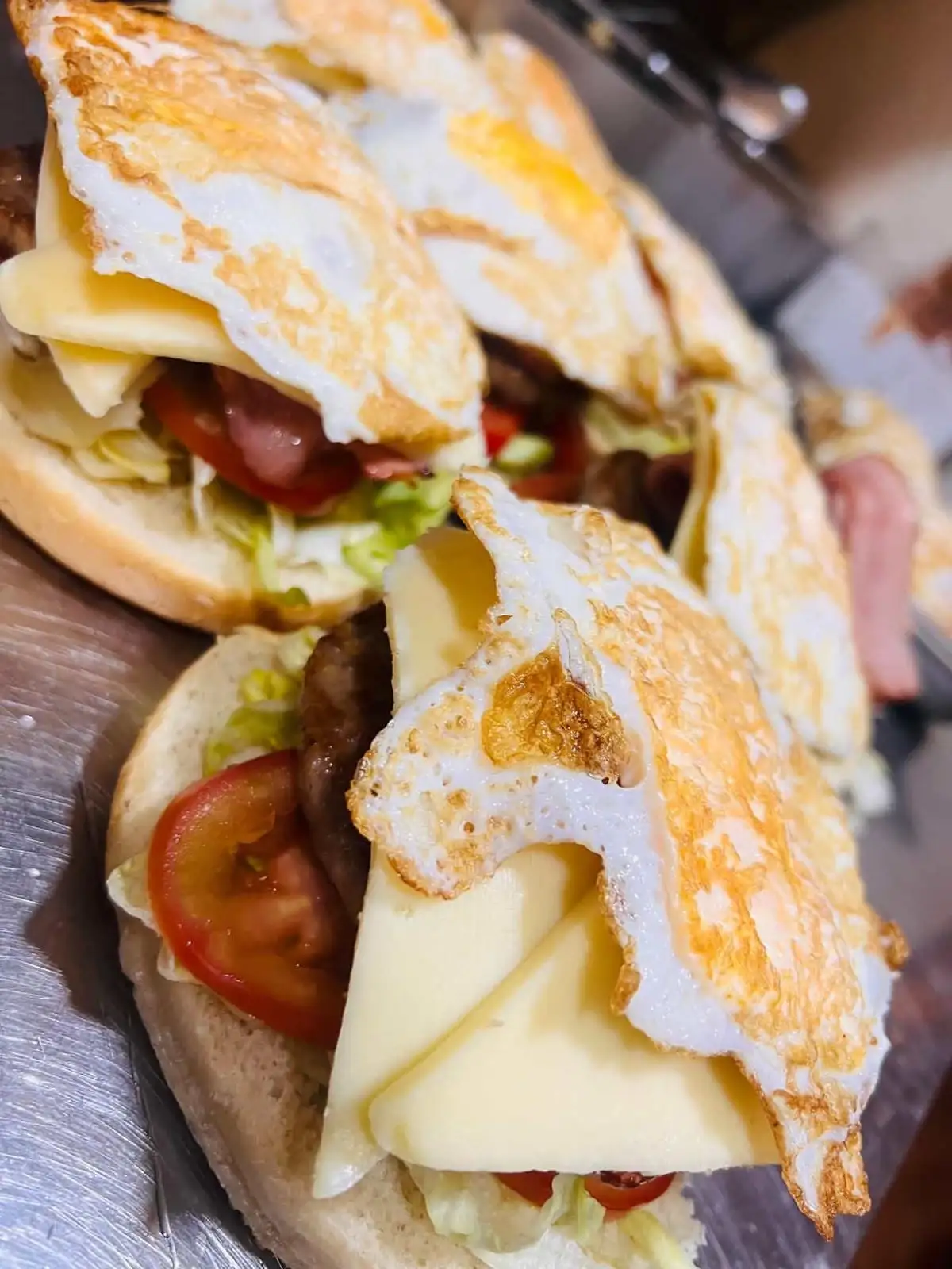 Open round-bun sandwiches with lettuce, tomato, grilled meat, slices of cheese and a fried egg crowning each one.