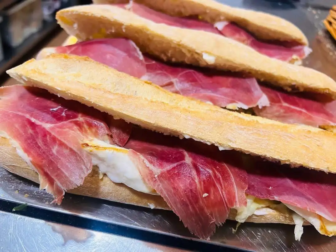 Baguette sandwiches lined up on a tray, open and covered with thin slices of serrano ham.