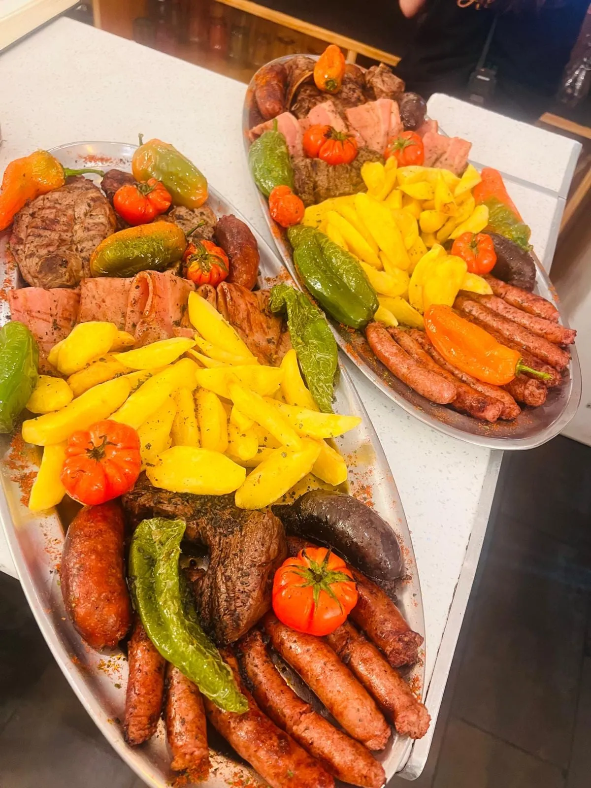 Two oval mixed-grill platters served in the dining room: grilled steaks and meat, blood sausage, pork sausages, potato wedges, green peppers and roasted tomato.