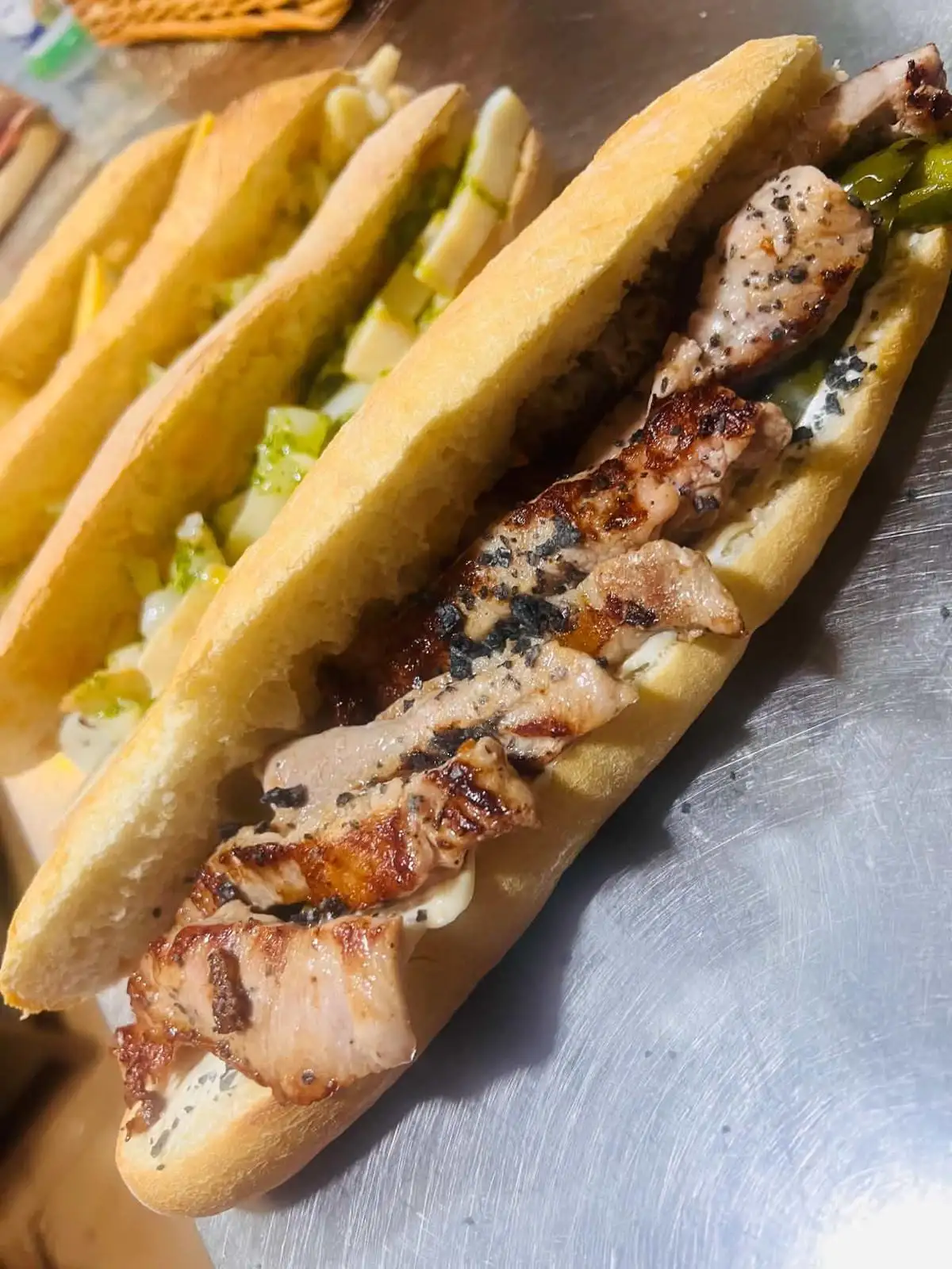 A baguette sandwich with grilled pork loin and black pepper, with two more behind filled with lettuce and cheese.