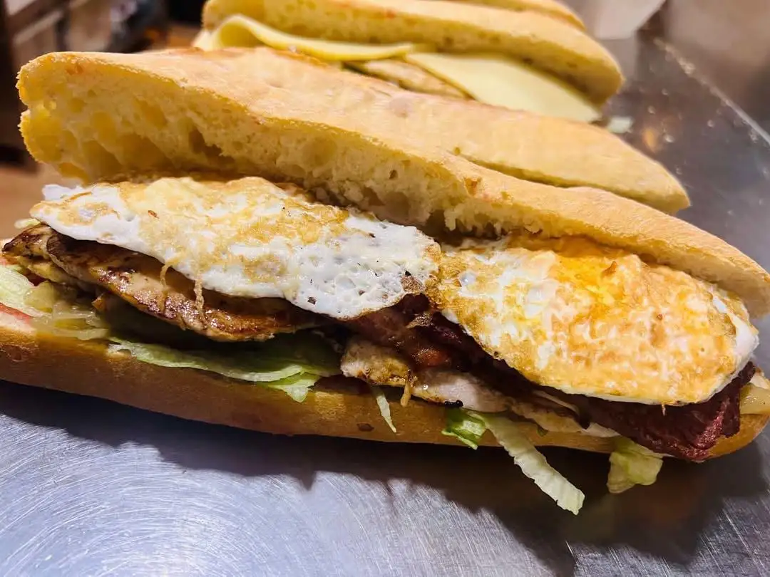 A ciabatta sandwich with lettuce, grilled pork loin, a strip of red sausage and a fried egg, with a cheese one behind.