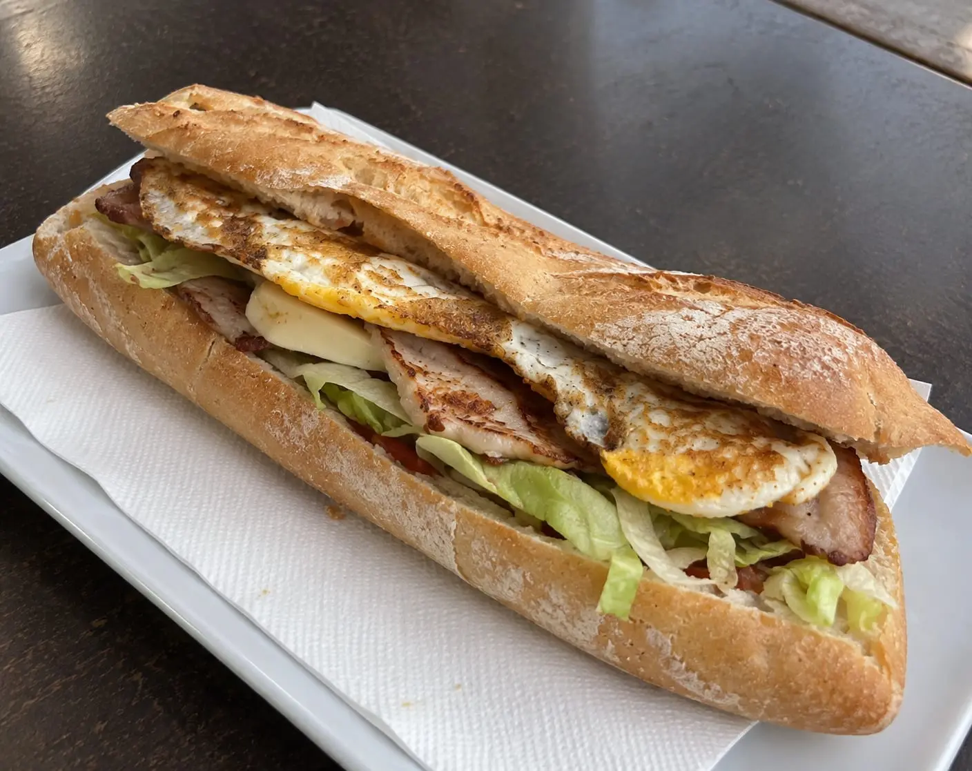 Entrepà de barra llarga amb llom, formatge, encisam i ou ferrat, servit en un plat blanc.