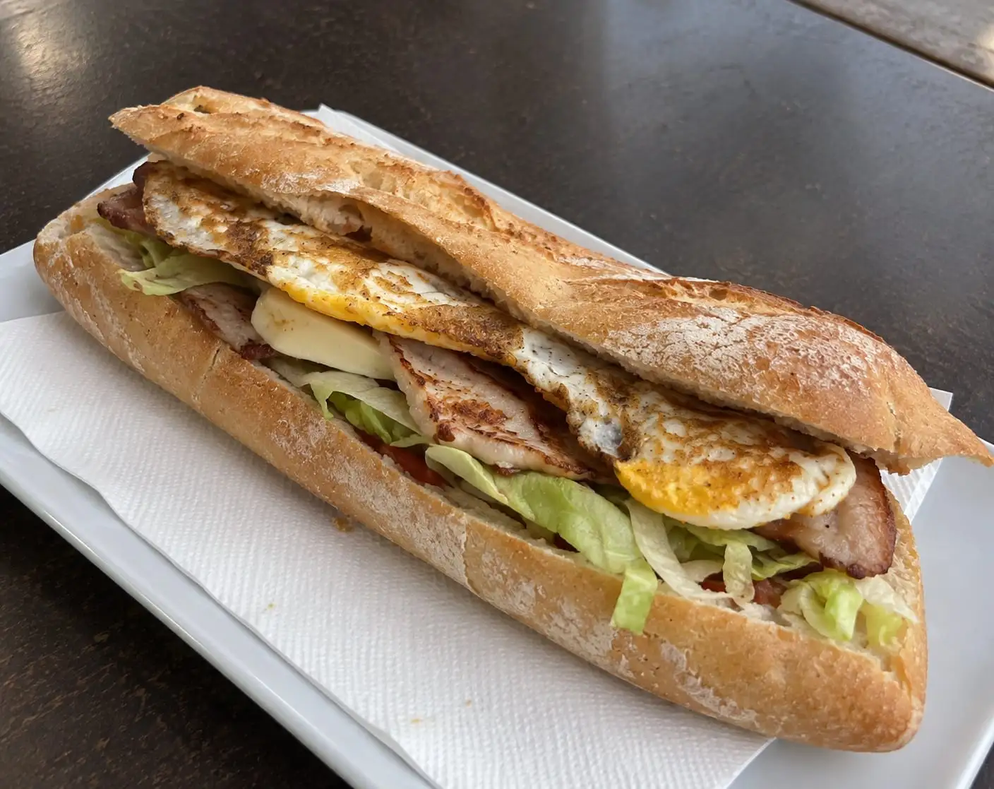 Long baguette roll with pork loin, cheese, lettuce and a fried egg, served on a white plate.