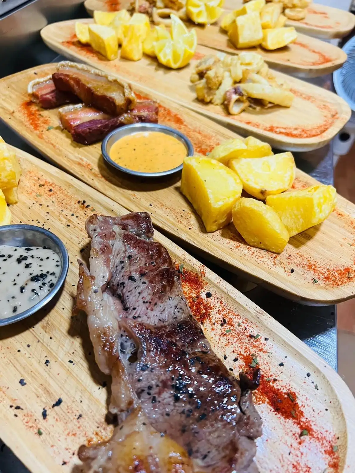 A row of wooden boards just served: rib steak, pork belly, squid, potatoes and carved lemon.