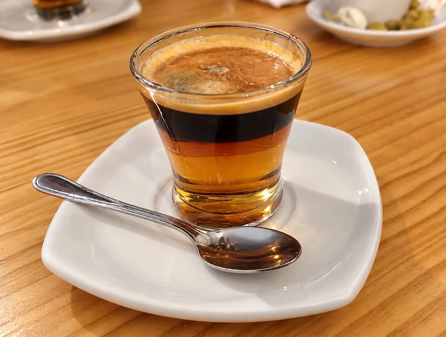 Coffee in a glass with its layers showing — condensed milk, coffee and crema — on a white saucer.