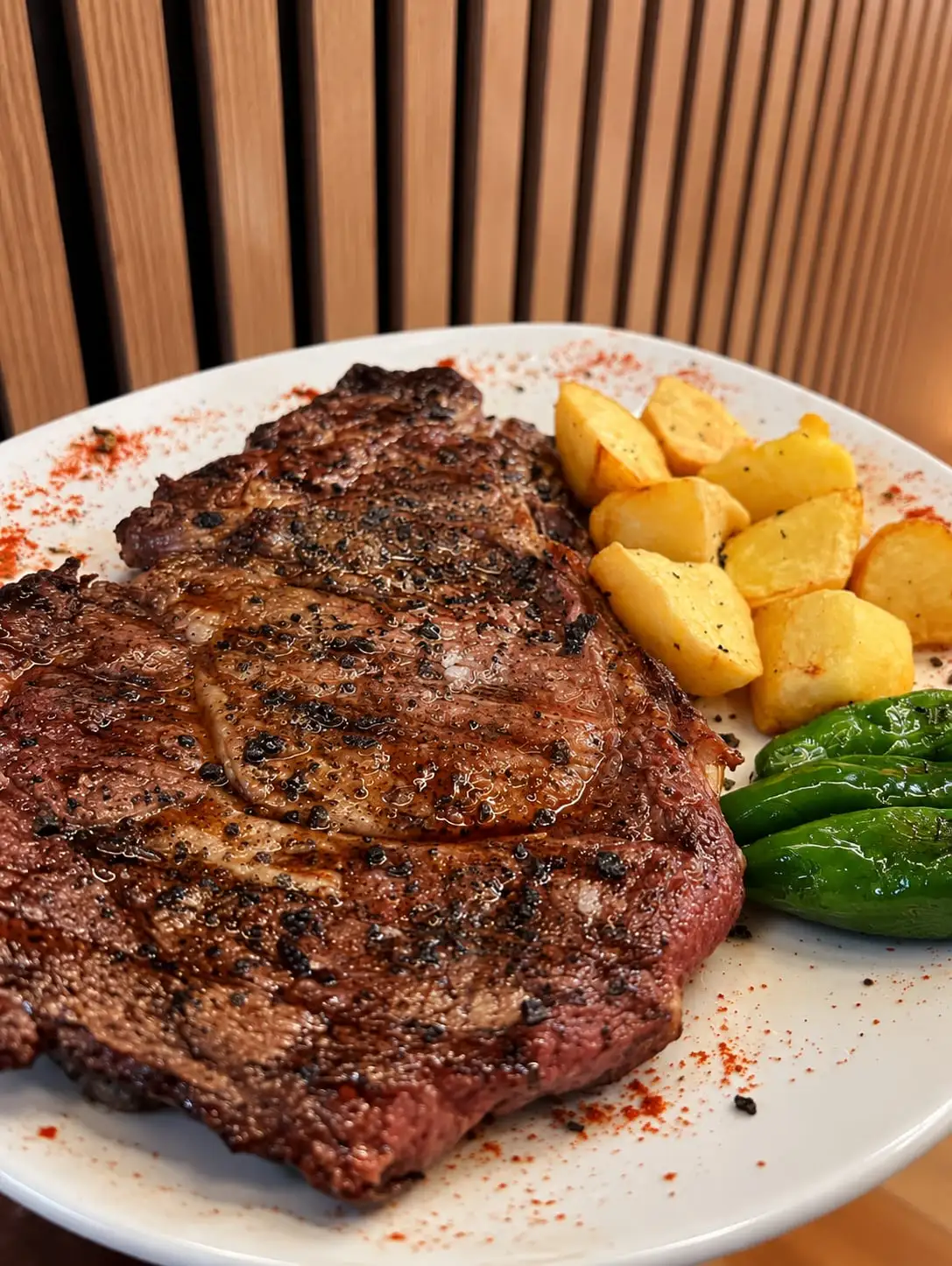Grilled rib steak on a white plate, with potatoes and green peppers beside it.