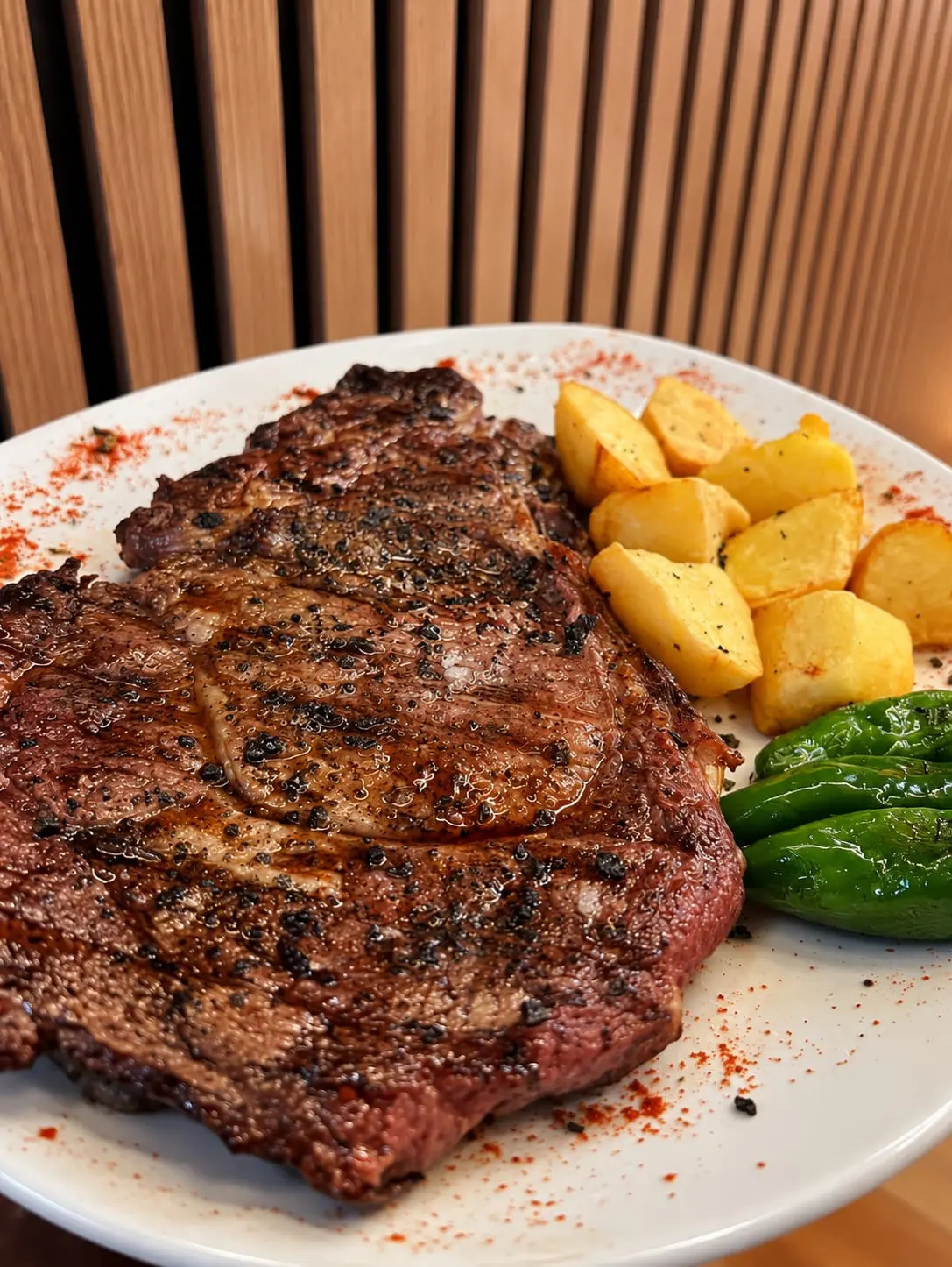 Bistec de costella fet a la brasa en un plat blanc, amb creïlles i pebrots verds al costat.